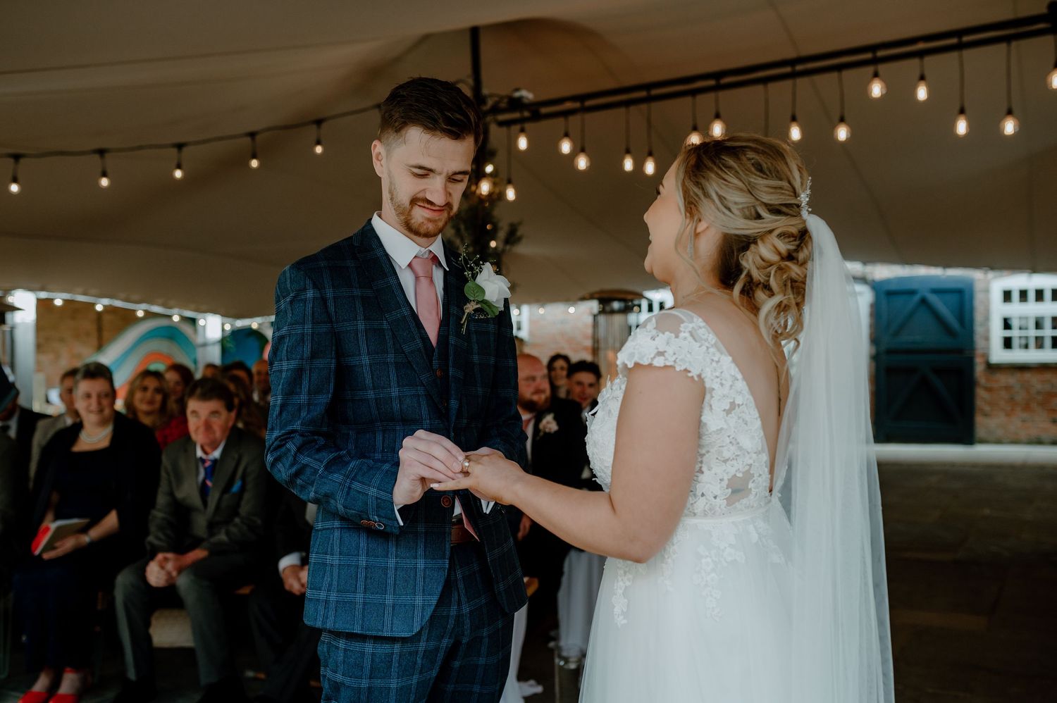 Couple exchanges rings during wedding ceremony under tent with string lights as guests watch in background.