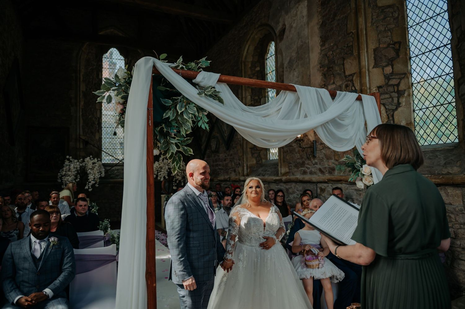 A wedding ceremony takes place inside a historic stone church with an elegant white draped arch and sunlit stained glass windows.