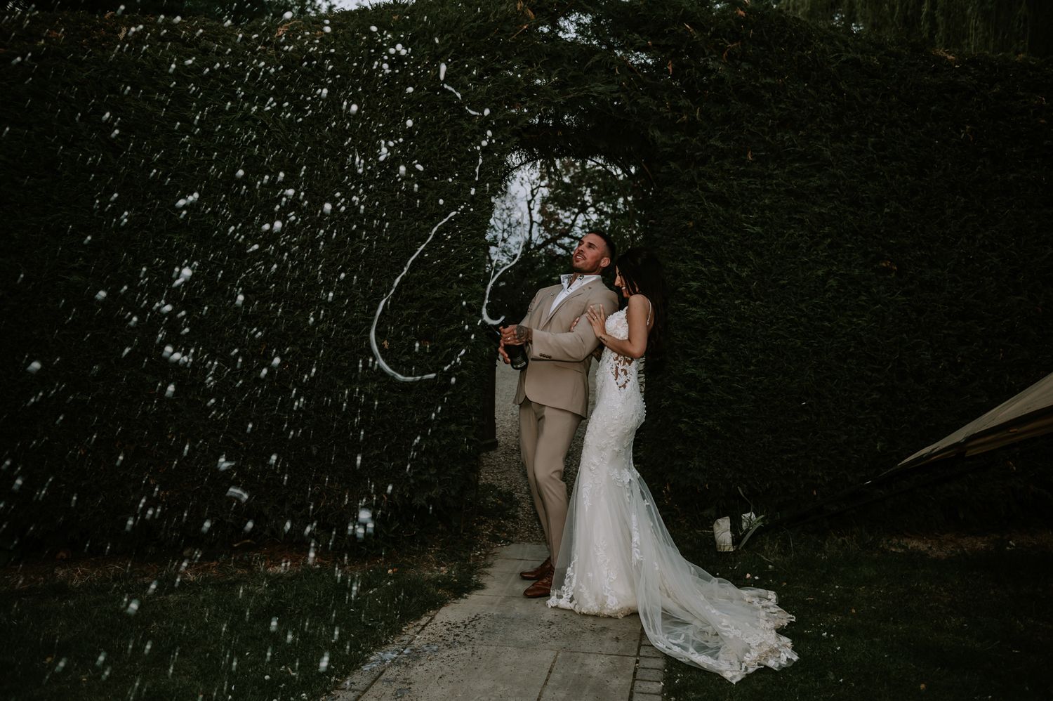 A couple shares an intimate moment in front of a lush green hedge while soap bubbles float around them.
