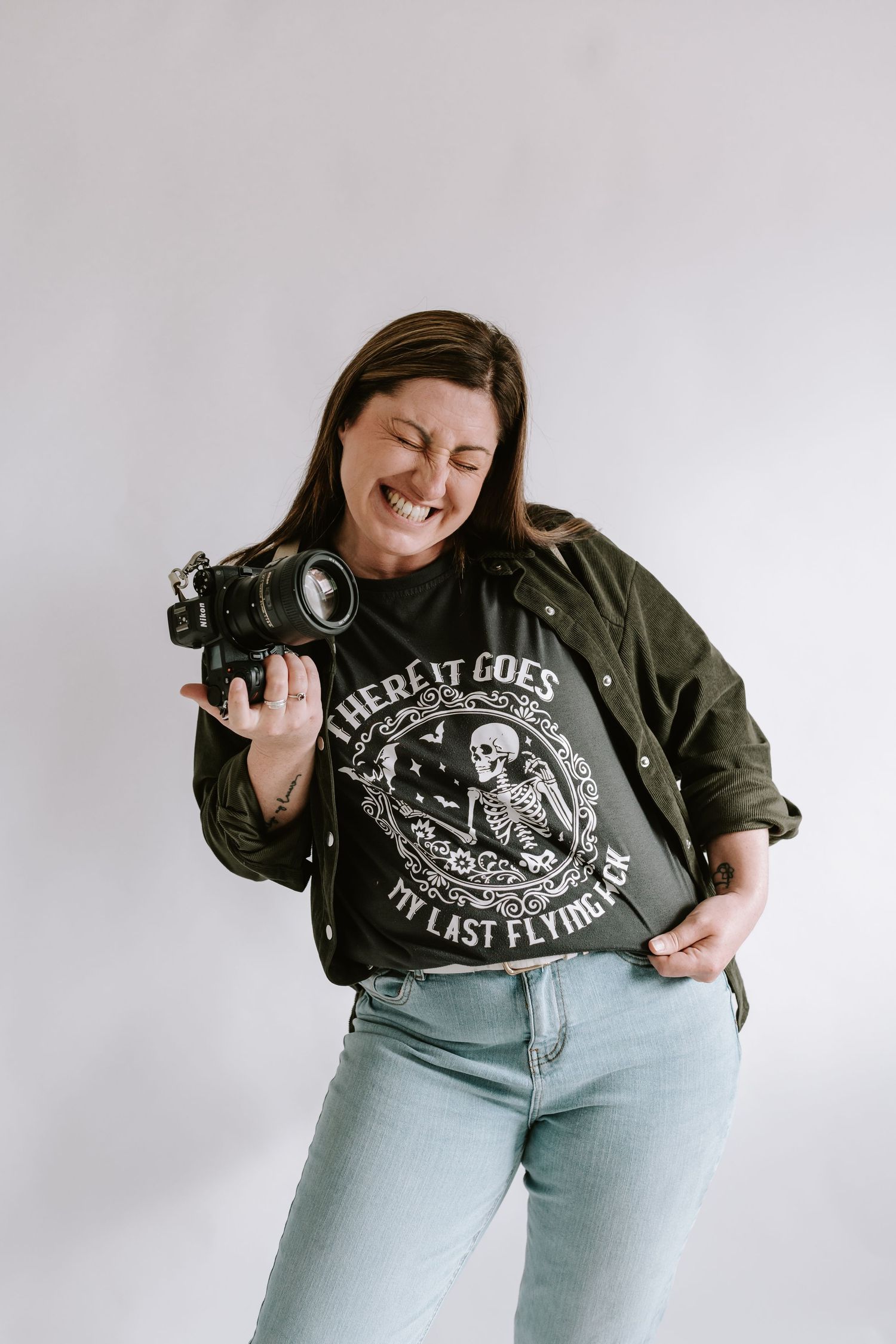Photographer wearing olive jacket and graphic t-shirt smiles while holding camera against grey background.