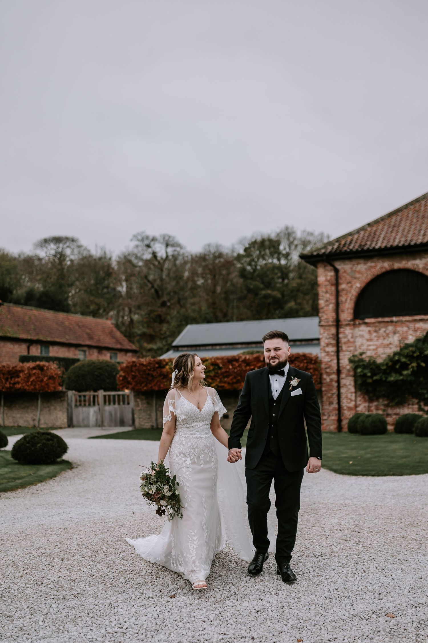 Newlywed couple holding hands on gravel path outside rustic brick venue with autumn garden and cloudy sky.