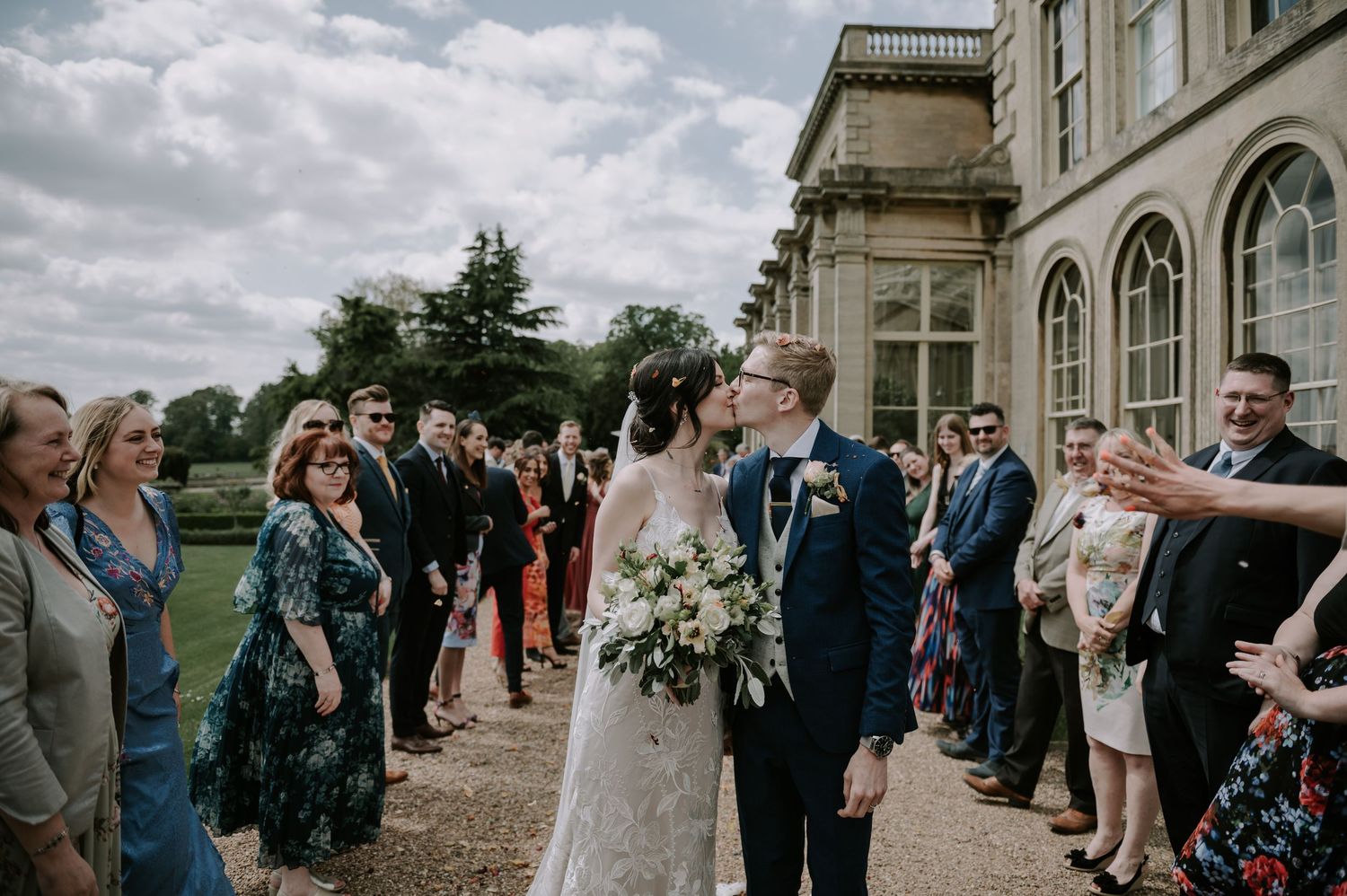Wedding guests line up outside an elegant stone building with arched windows as a couple shares a moment during their celebration.