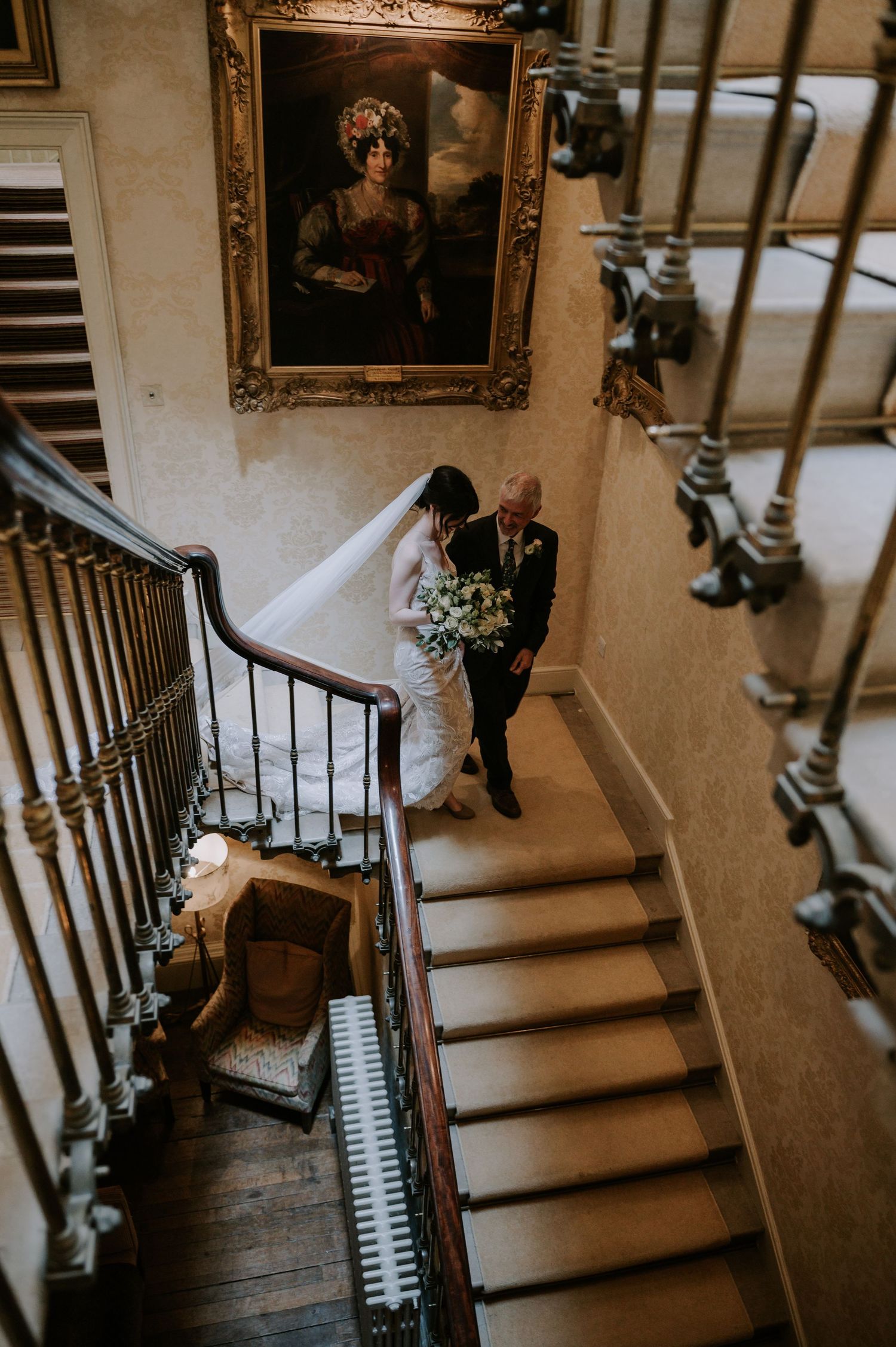 A graceful view down an elegant wooden staircase with vintage decor shows a wedding dress descending the steps.
