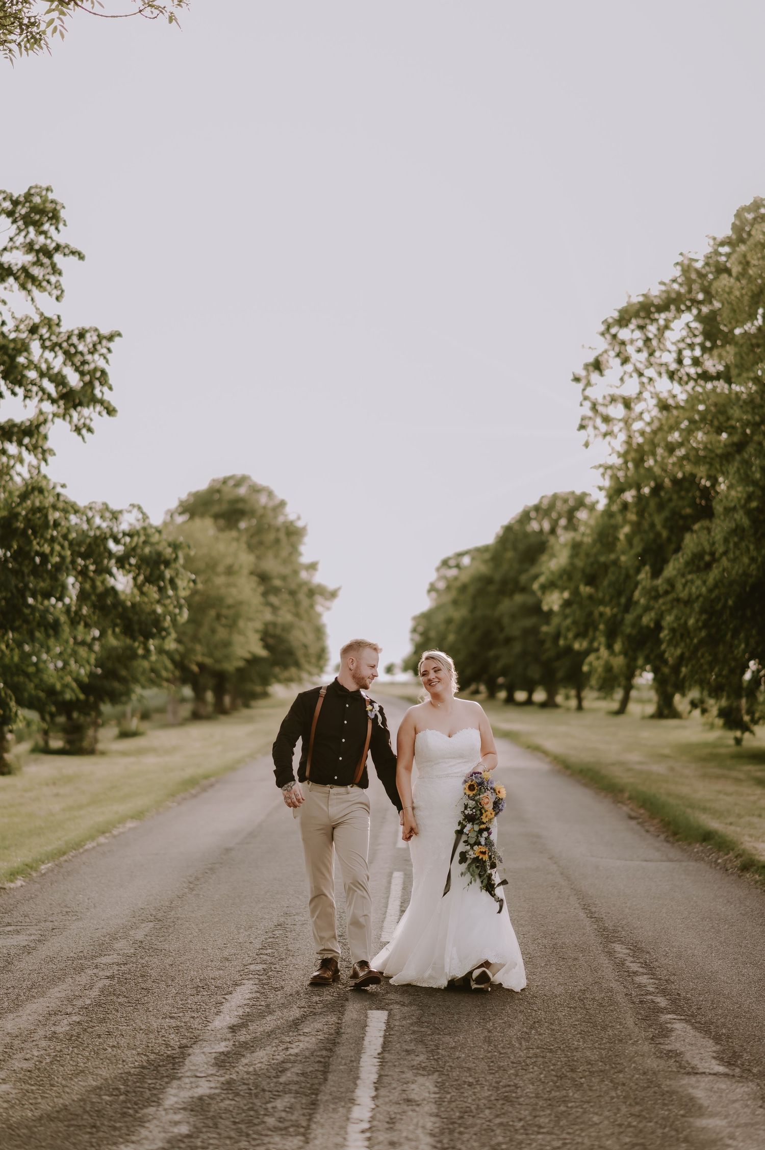 Bride and groom walk hand-in-hand down a tree-lined road, bride holding a bouquet in a white gown.