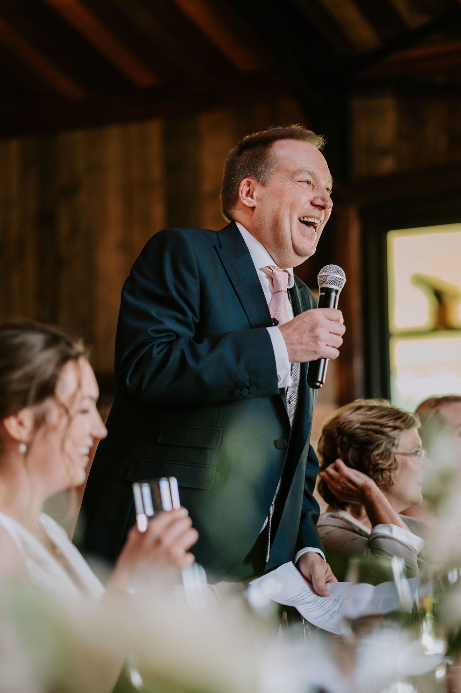 A speaker in a navy suit delivers a speech at a wedding reception, creating a lively and joyful atmosphere.