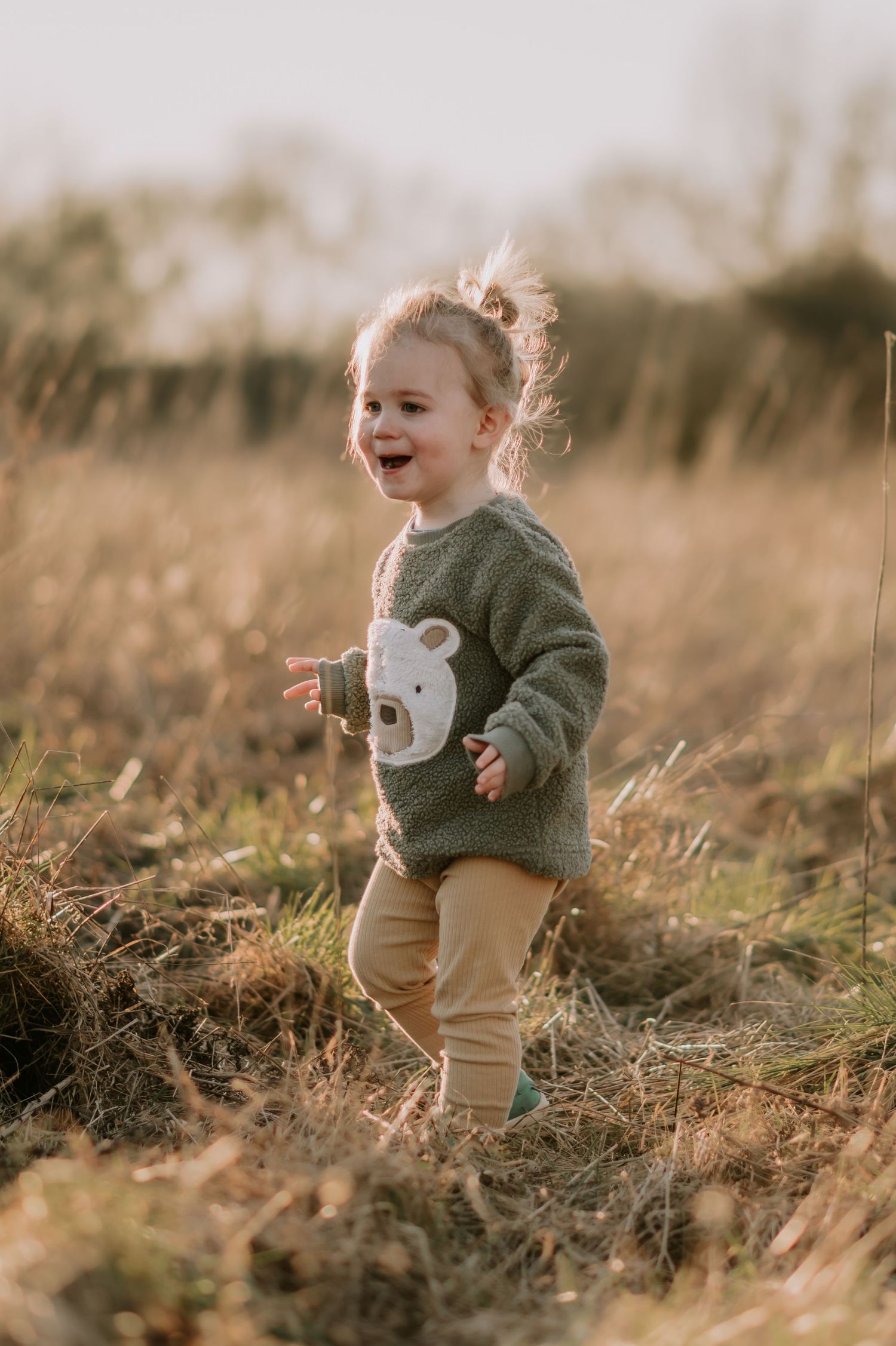 A child wearing a green sweater and beige pants plays joyfully in a golden field at sunset.