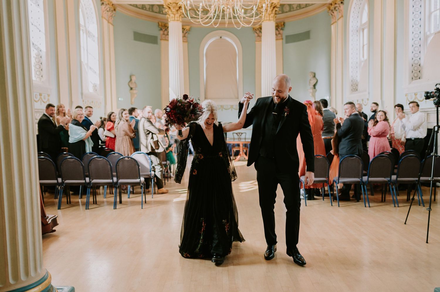 Two elegantly dressed figures in black walk hand in hand inside an ornate ballroom with columns and chandeliers.
