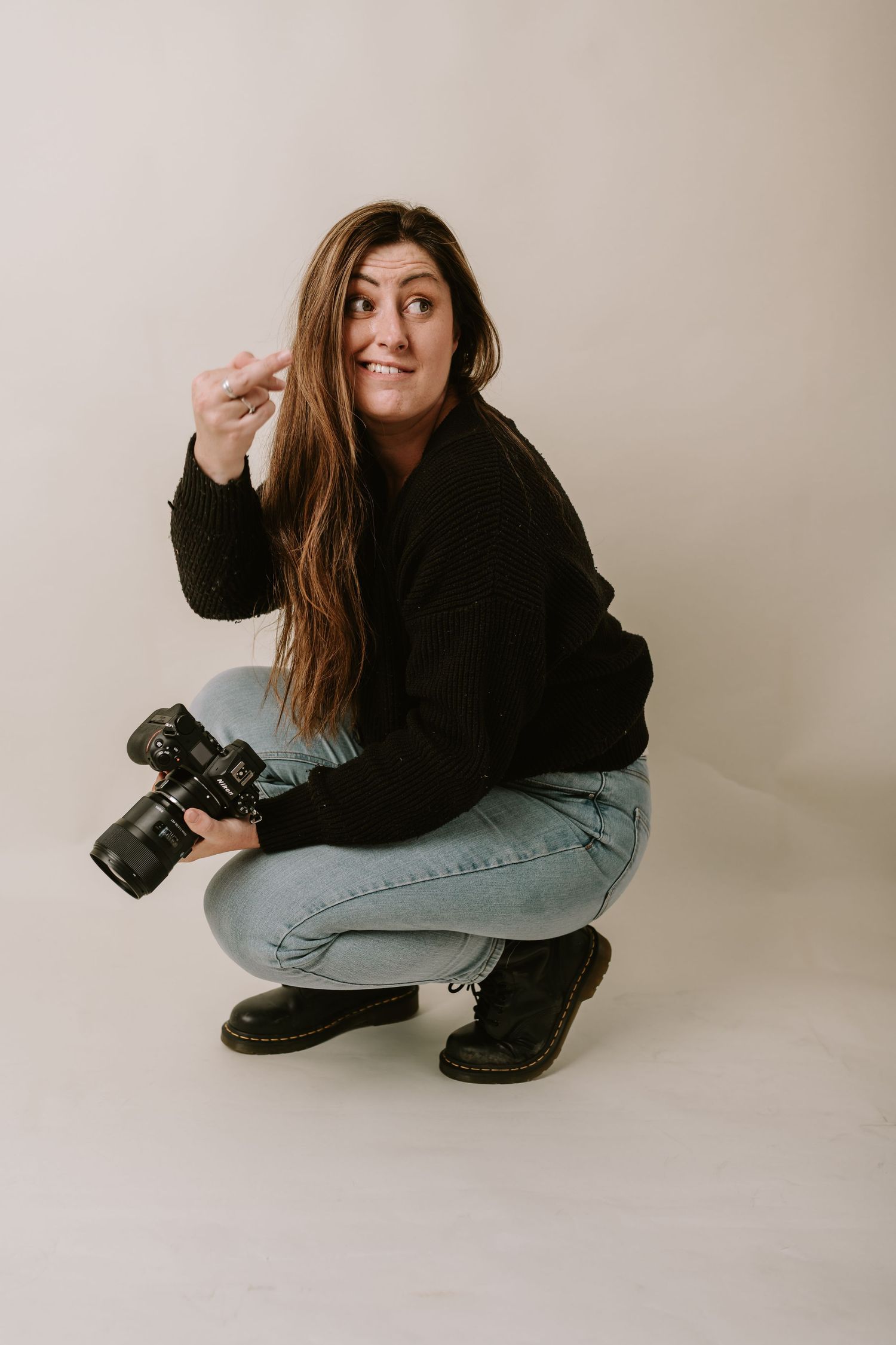 Professional photographer in black sweater and jeans crouches with camera while making playful expression in studio setting.