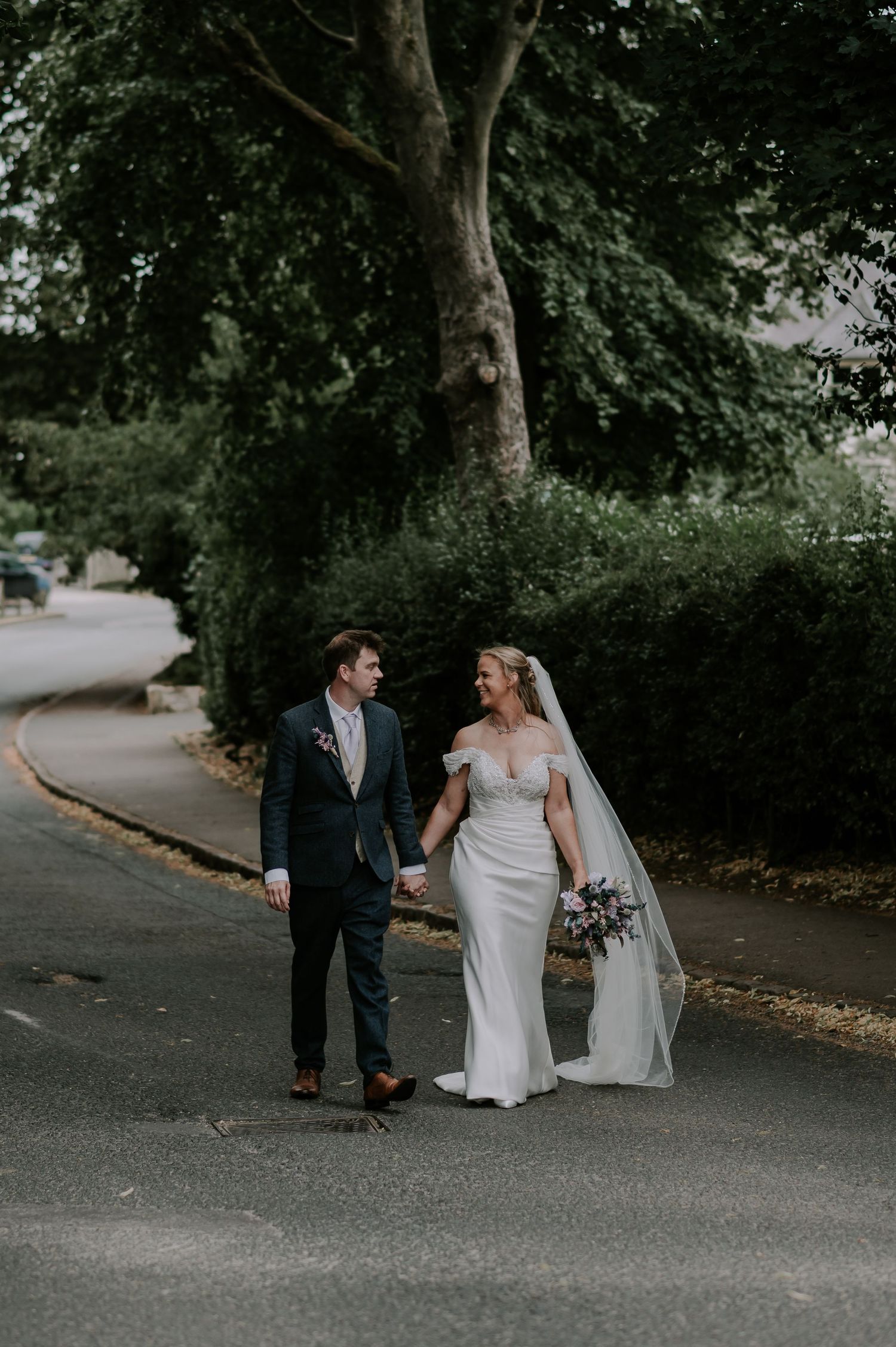 A couple walks together on a tree-lined path, with the bride wearing a fitted white gown and holding a bouquet.