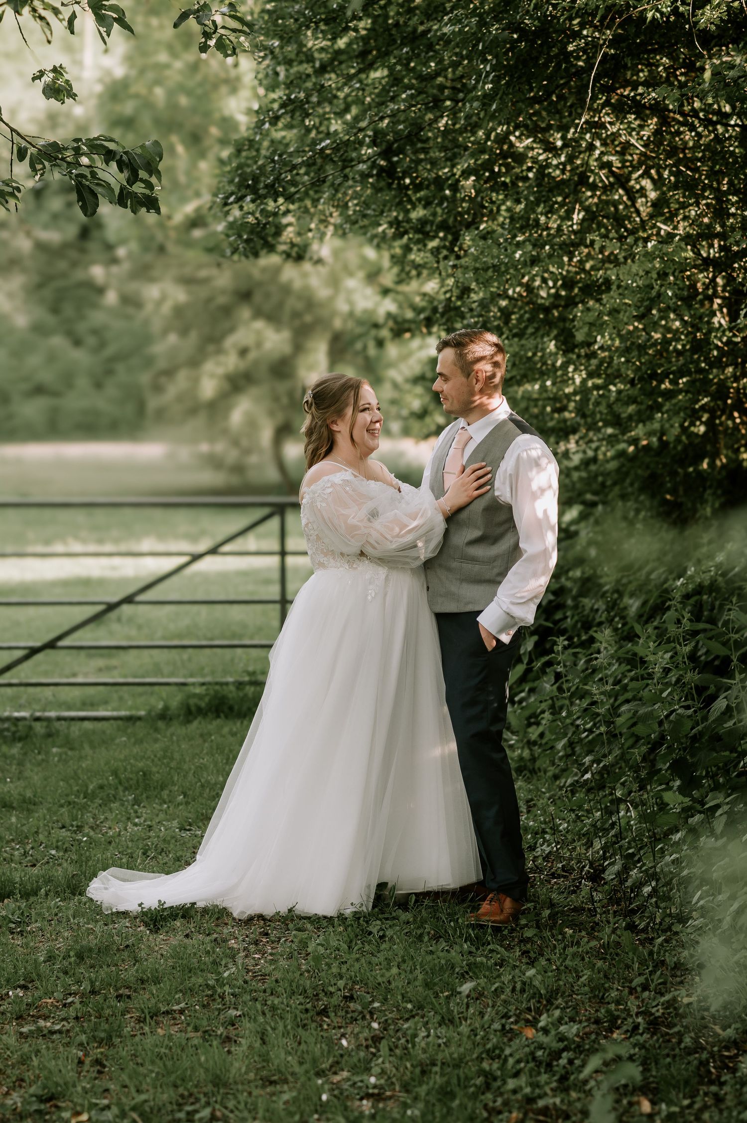 Couple shares romantic moment in white wedding attire against lush garden backdrop with metal gate.