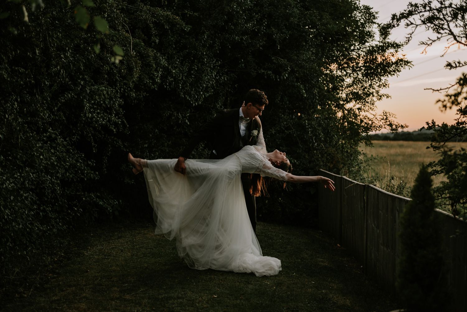 A romantic sunset photography pose as a couple creates a dramatic lean against a fence overlooking a countryside field.