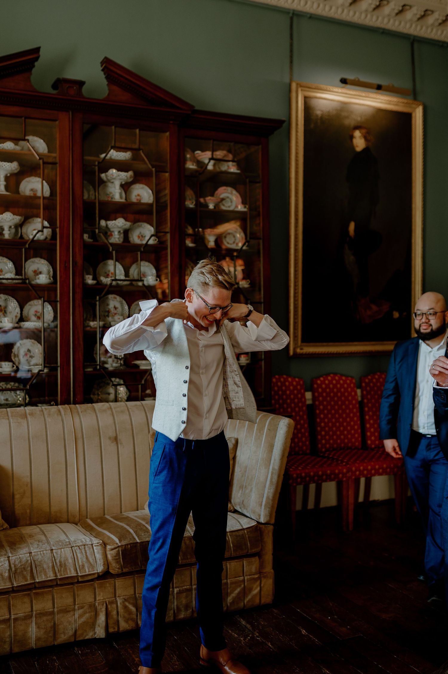 Two people in a room with antique display cabinets filled with china and a portrait painting on the mint green wall.