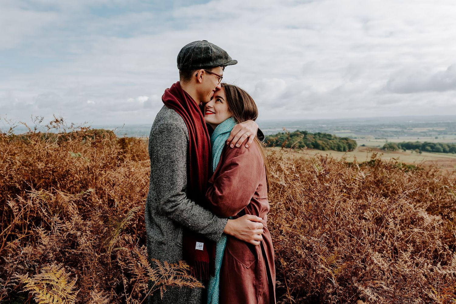 A couple embraces in warm clothing while standing among autumn foliage on a hilltop with cloudy skies in the background.