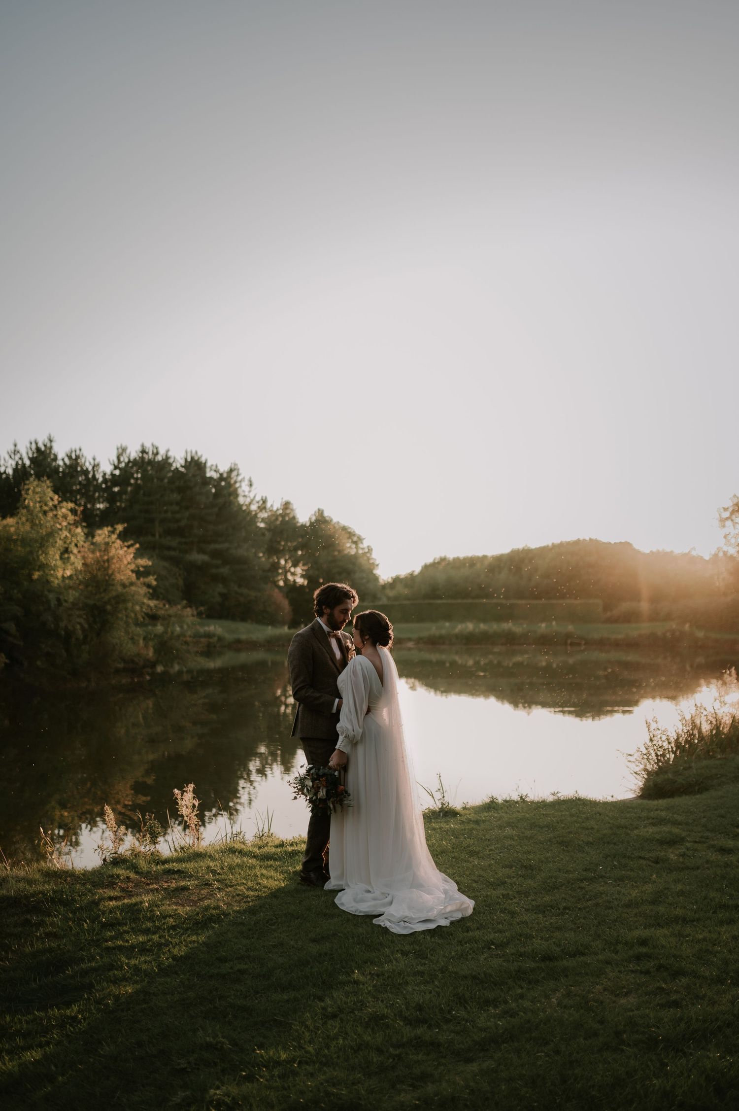 Newlywed couple stands together by a serene lake at sunset creating romantic silhouettes against the golden sky.