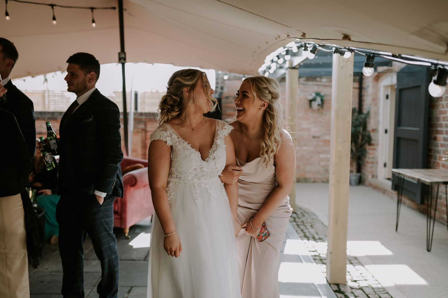 A bride and bridesmaid share a candid moment in an elegant indoor wedding venue with string lights overhead.