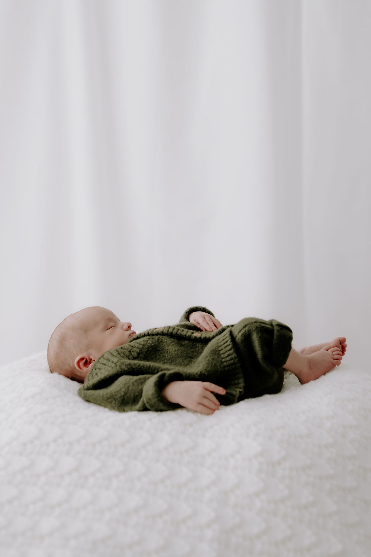 Newborn baby in green knit sweater lying on white bedding in studio – Melton Mowbray newborn photoshoot.