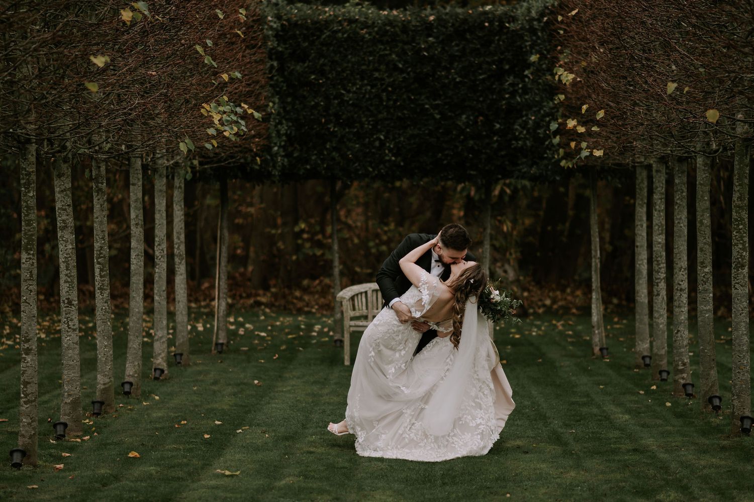 Couple sharing dip kiss on lush garden path lined with tall trees during autumn – Hazel Gap Barn, Derby wedding.