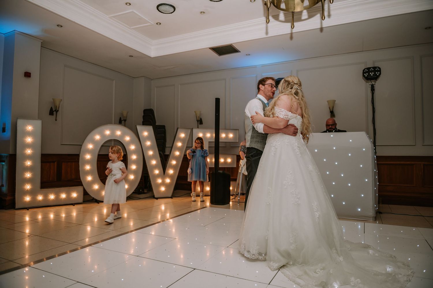 A couple shares their first dance on an illuminated dance floor with glowing LOVE letters in the background at their wedding reception.
