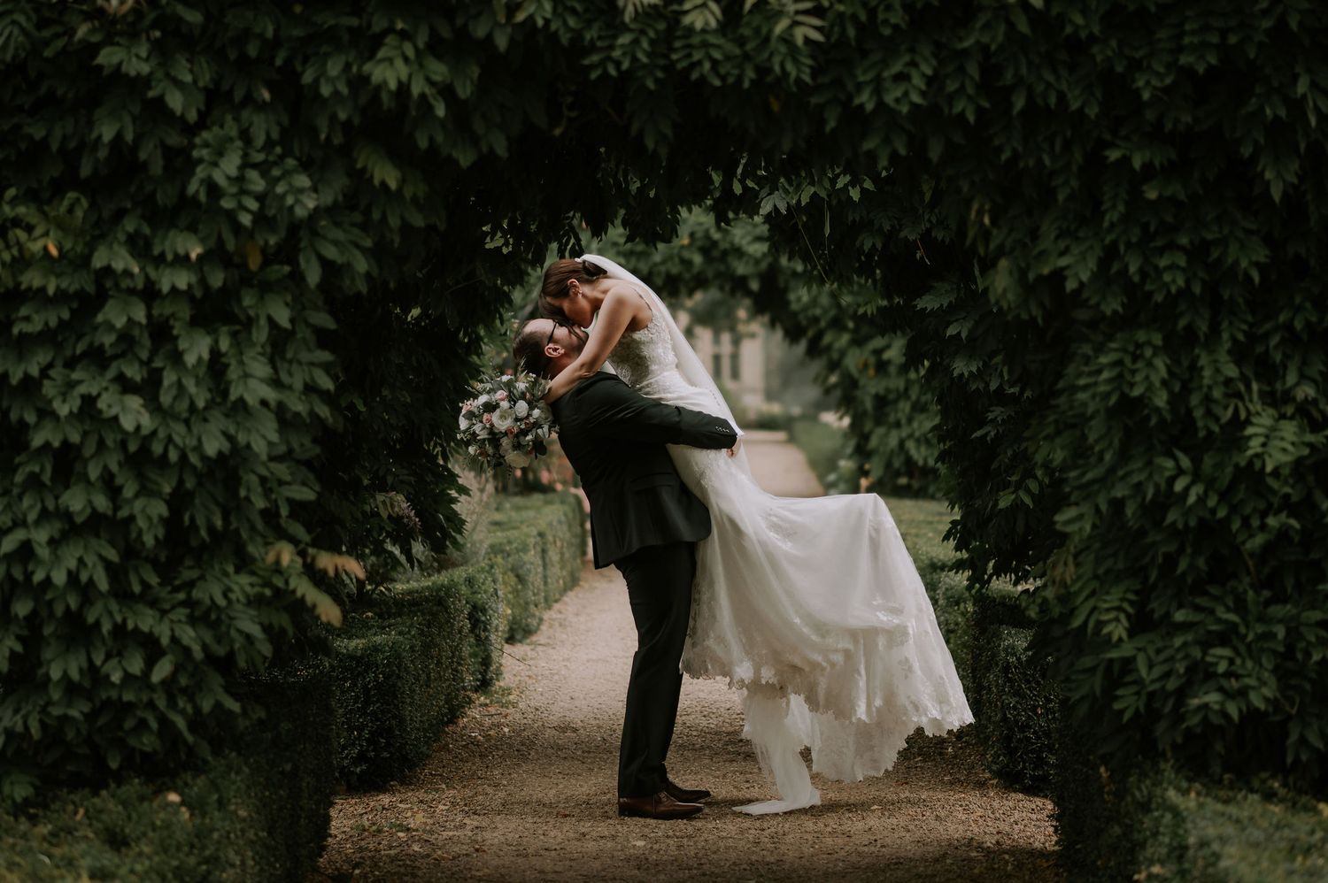 Groom lifts up his new bride under an archway of greenery in a romantic kiss.