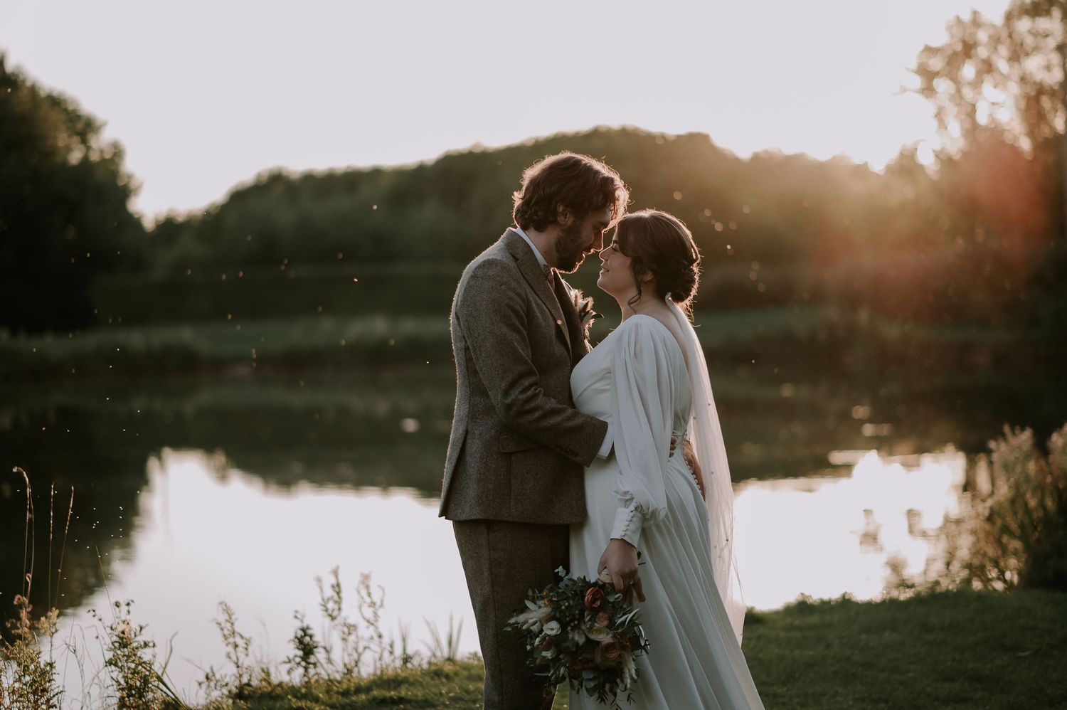 Couple sharing romantic sunset moment by serene lake with floral bouquet – Hidden Hive, Swadlincote wedding.