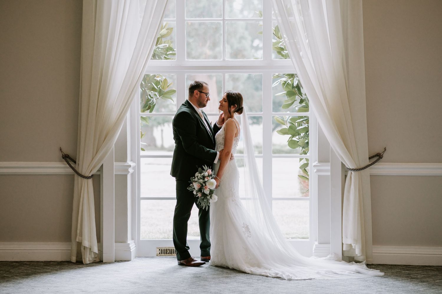 A romantic wedding moment silhouetted by natural light streaming through tall white-curtained windows.