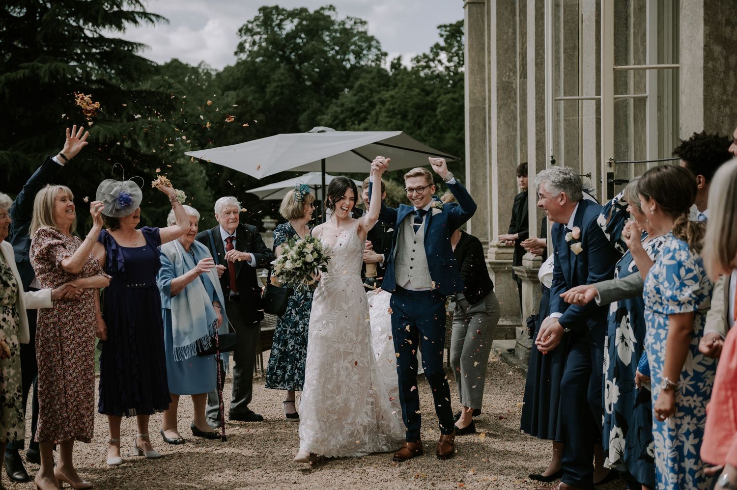 Wedding celebration outside historic building as guests throw confetti while couple exits through stone columns.