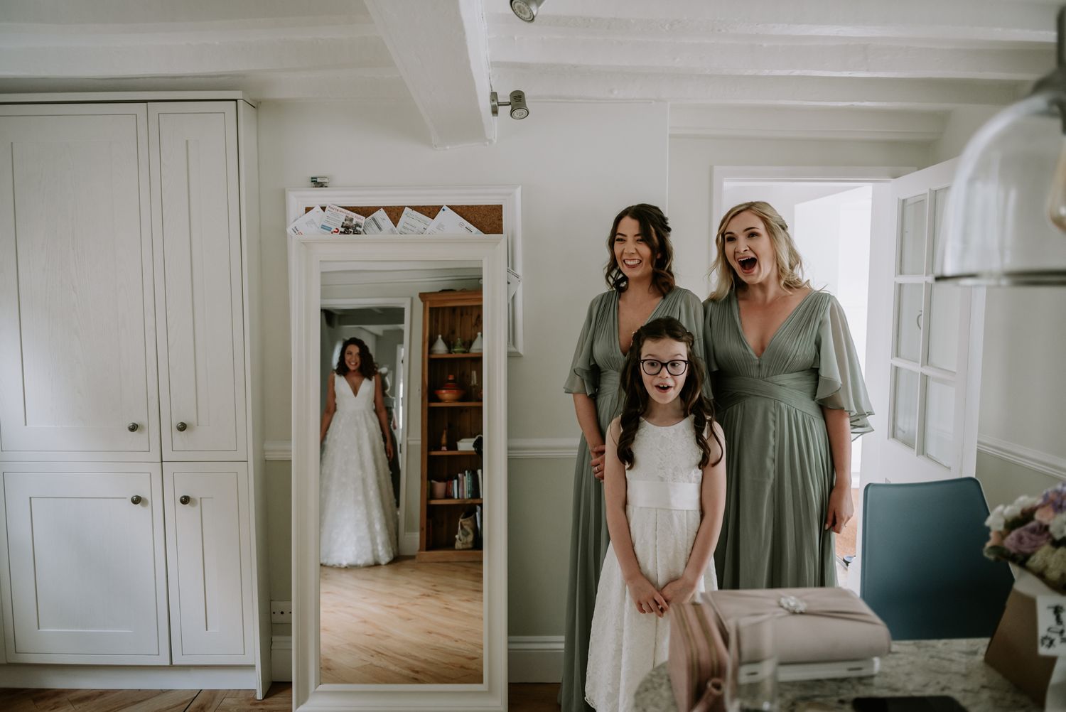 Three people stand in front of a mirror during a wedding preparation in a white dressing room.
