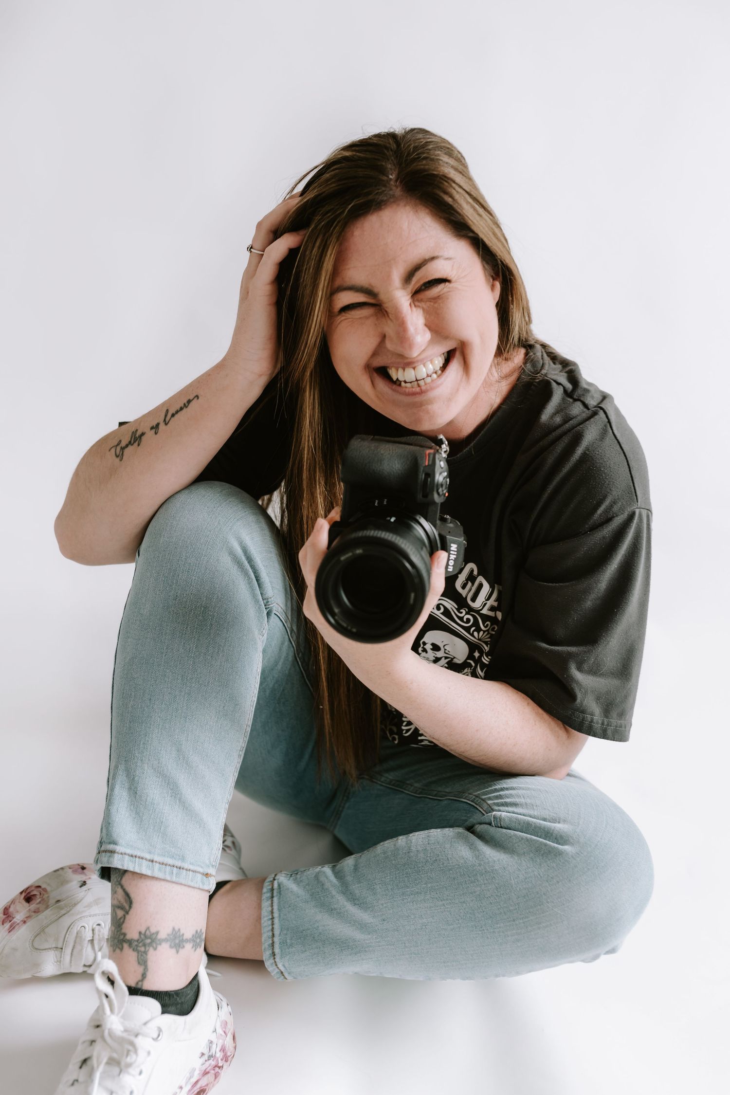 Creative photographer sitting cross-legged with camera and laughing joyfully against white backdrop.