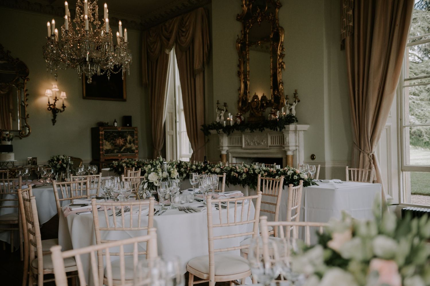 Elegant wedding reception tables set with crystal chandeliers, white chairs, and floral centerpieces in a bright venue.
