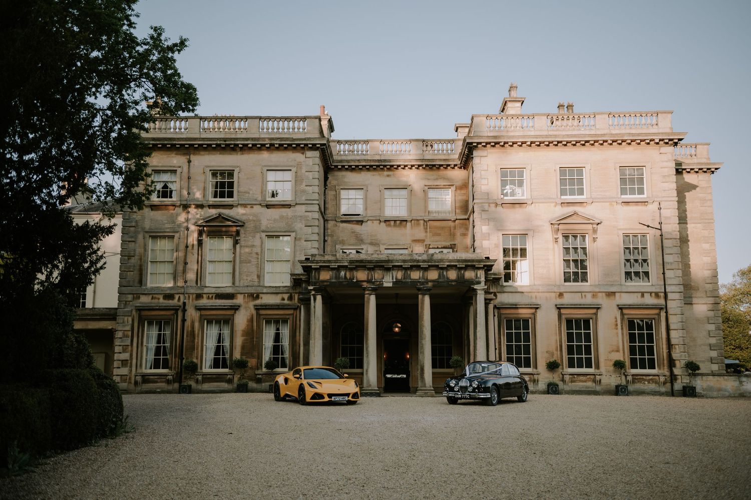 A stately stone mansion with classic Georgian architecture and vintage cars parked in front of its grand entrance.