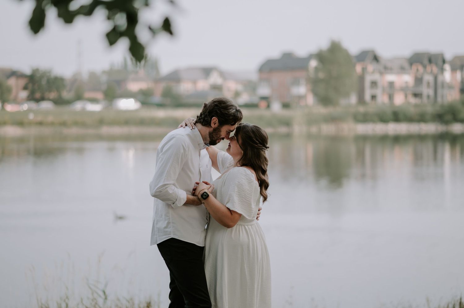 A couple in white sharing a romantic moment by a serene lake with houses visible in the background.