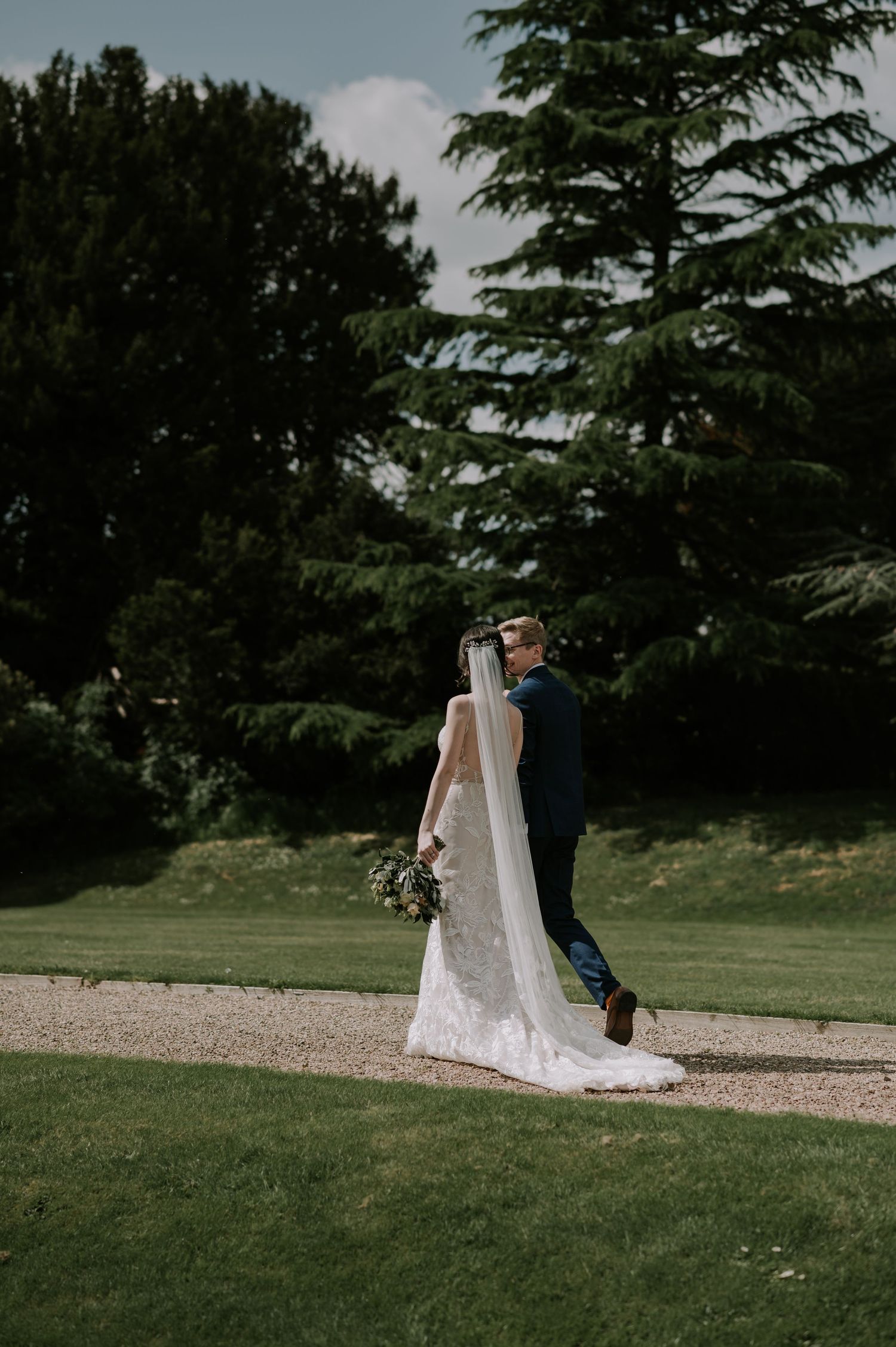 Elegant wedding couple shares intimate moment walking down gravel path surrounded by lush green trees and lawn.