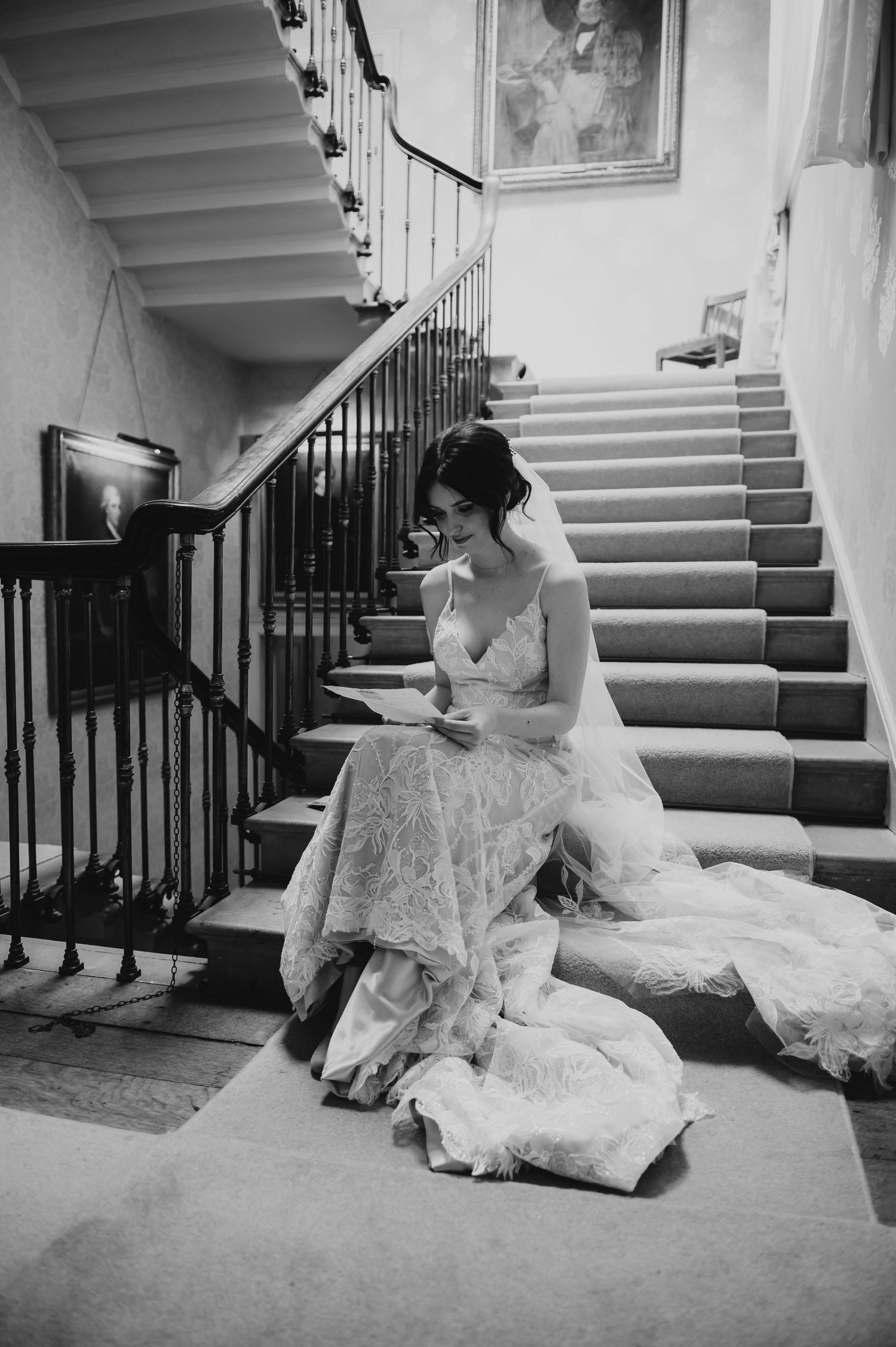 A person in an elegant lace wedding gown reads while seated on grand carpeted stairs in a classic black and white photo.