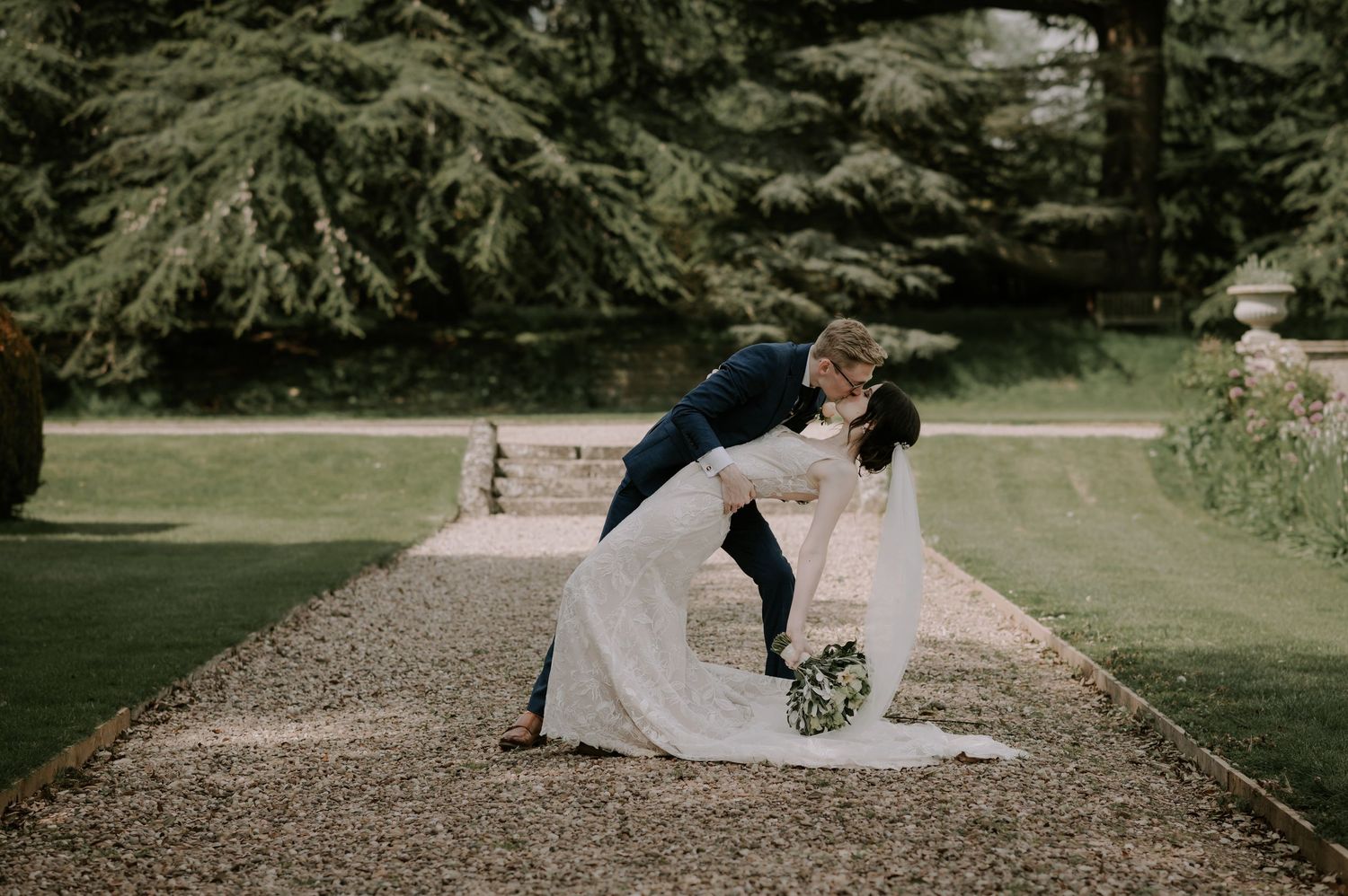 A couple in elegant wedding attire share an intimate moment against a backdrop of lush greenery in a garden setting.