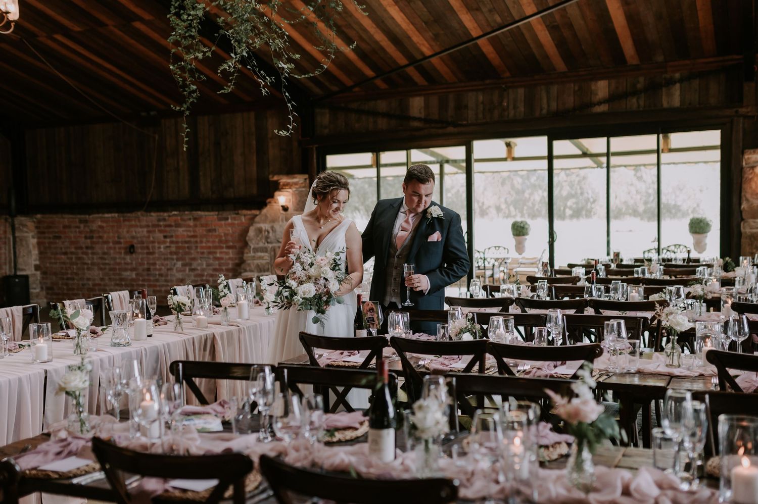 Elegant wedding reception venue with wooden beams, exposed brick walls, and tables set with white linens and centerpieces.