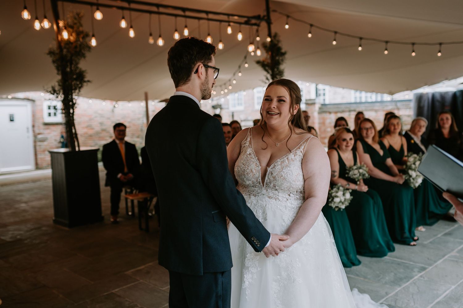 A beautiful wedding ceremony takes place under a tent with string lights and emerald-dressed bridesmaids seated in the background.