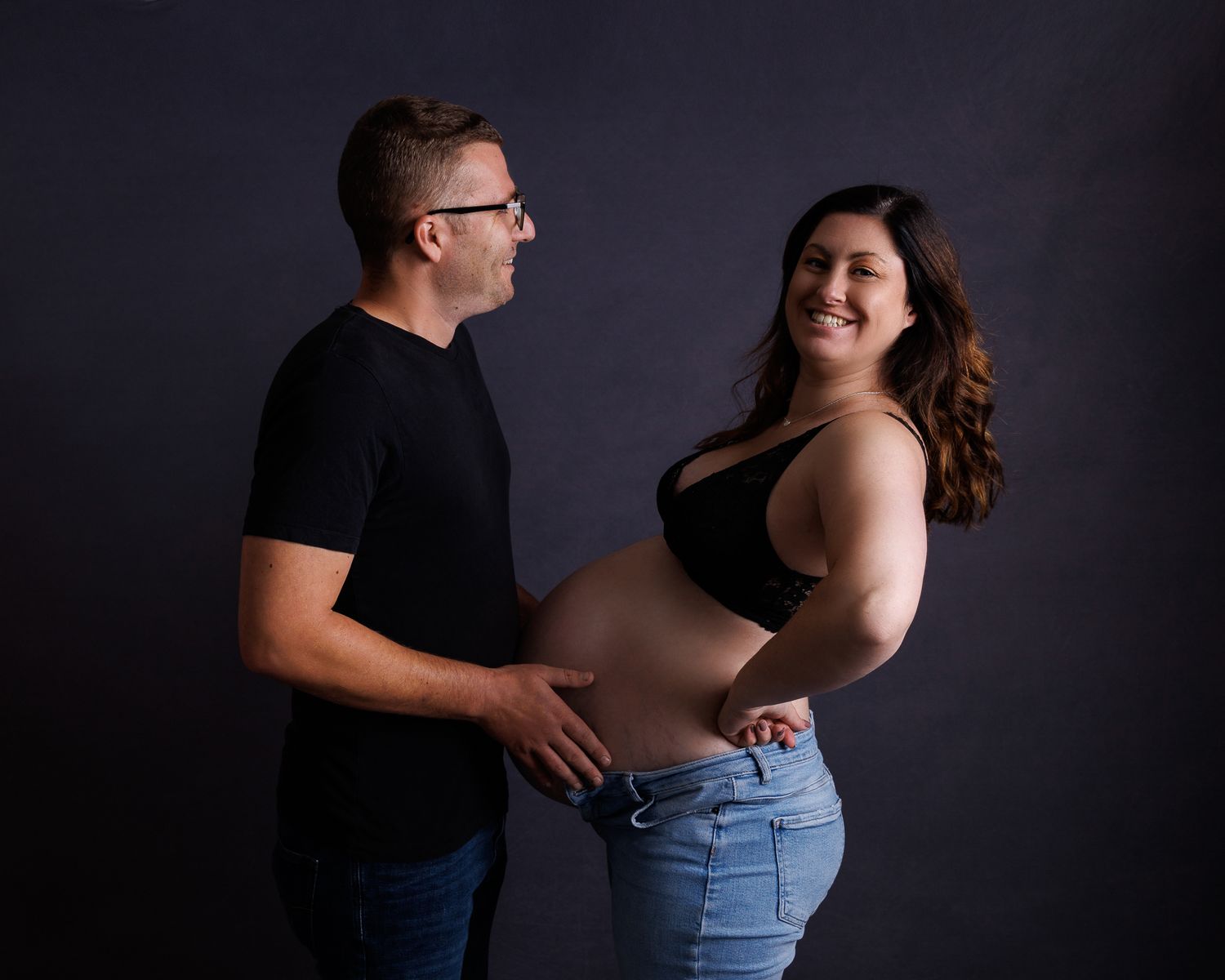Heavily pregnant woman playfully pressing her baby bump against her husband while smiling, studio maternity portrait with a dark background.