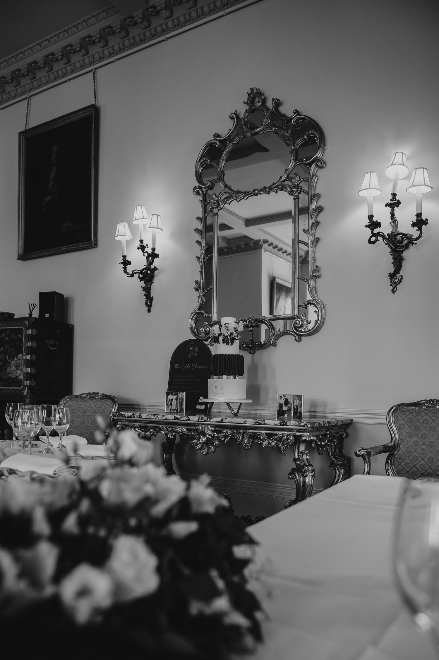 Elegant ballroom interior with ornate wall sconces and floral centerpieces on white tablecloths in vintage black and white style.