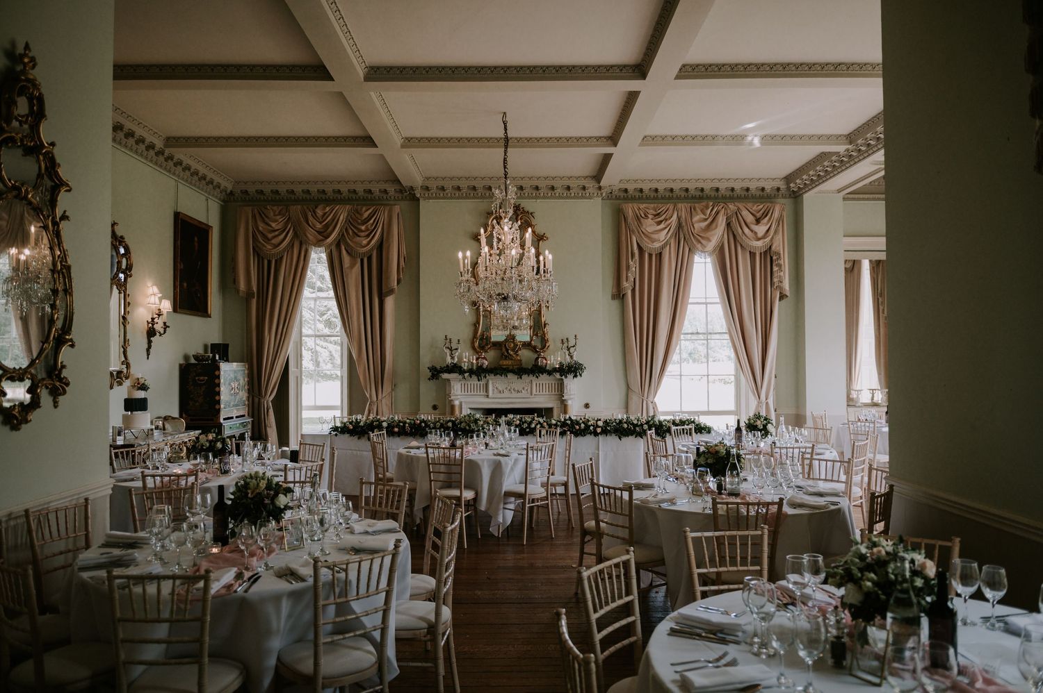 An elegant wedding reception ballroom with crystal chandeliers, round dining tables, and sage green walls.