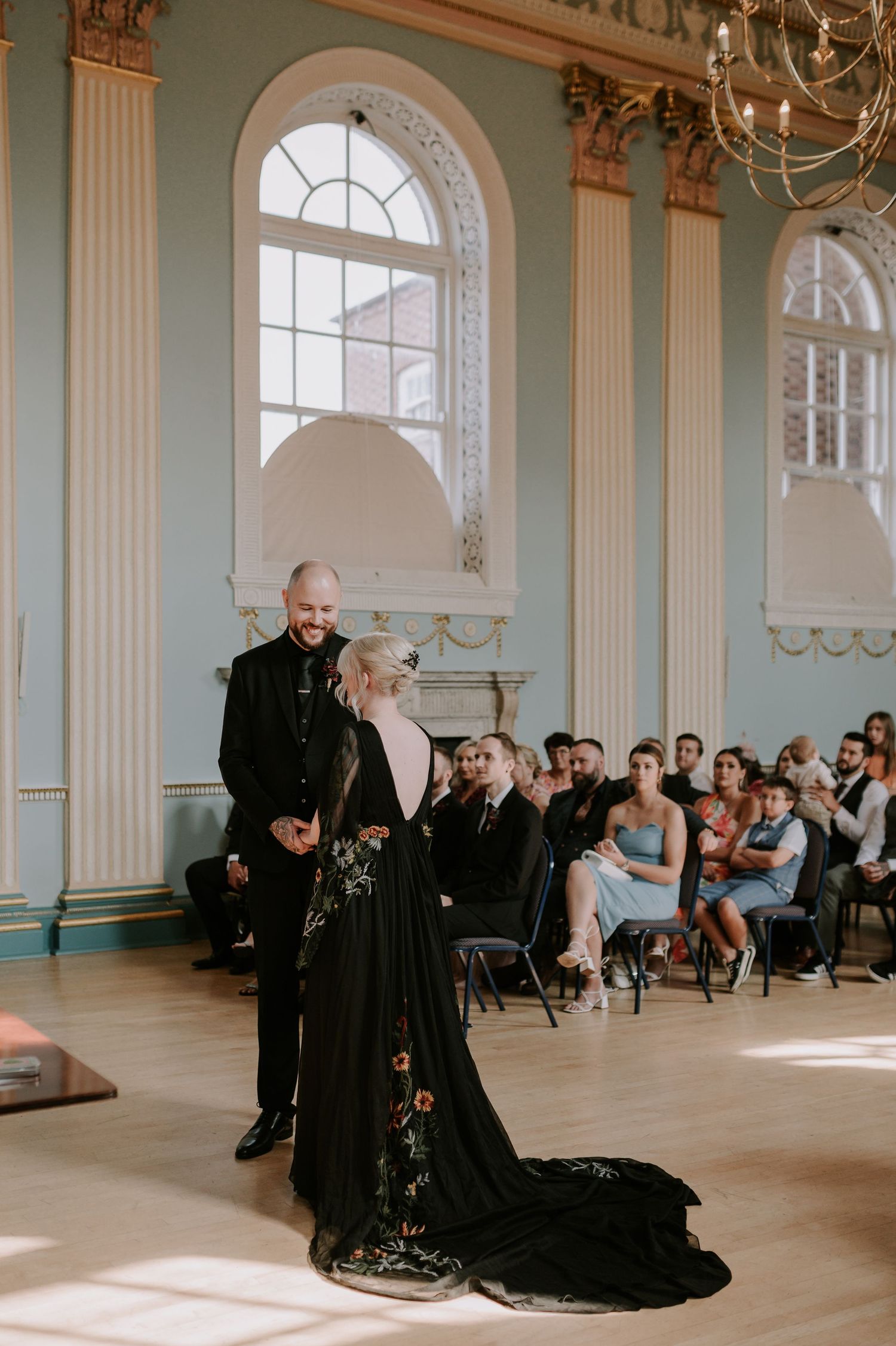 Elegant dancers in black attire perform a dramatic sequence across a grand ballroom with ornate columns and arched windows.
