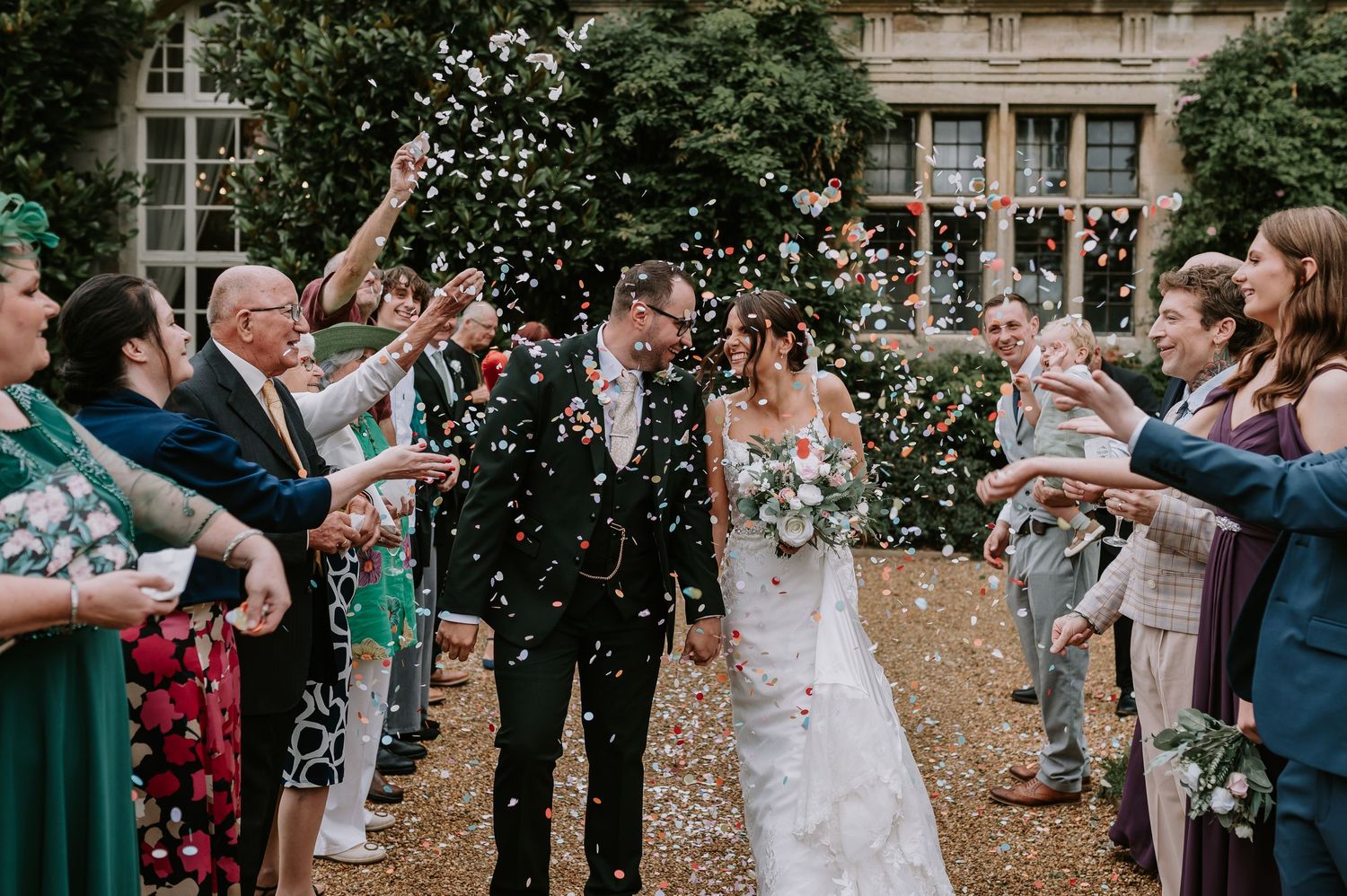 Wedding guests toss white confetti over a couple walking down an outdoor aisle lined with well-wishers.
