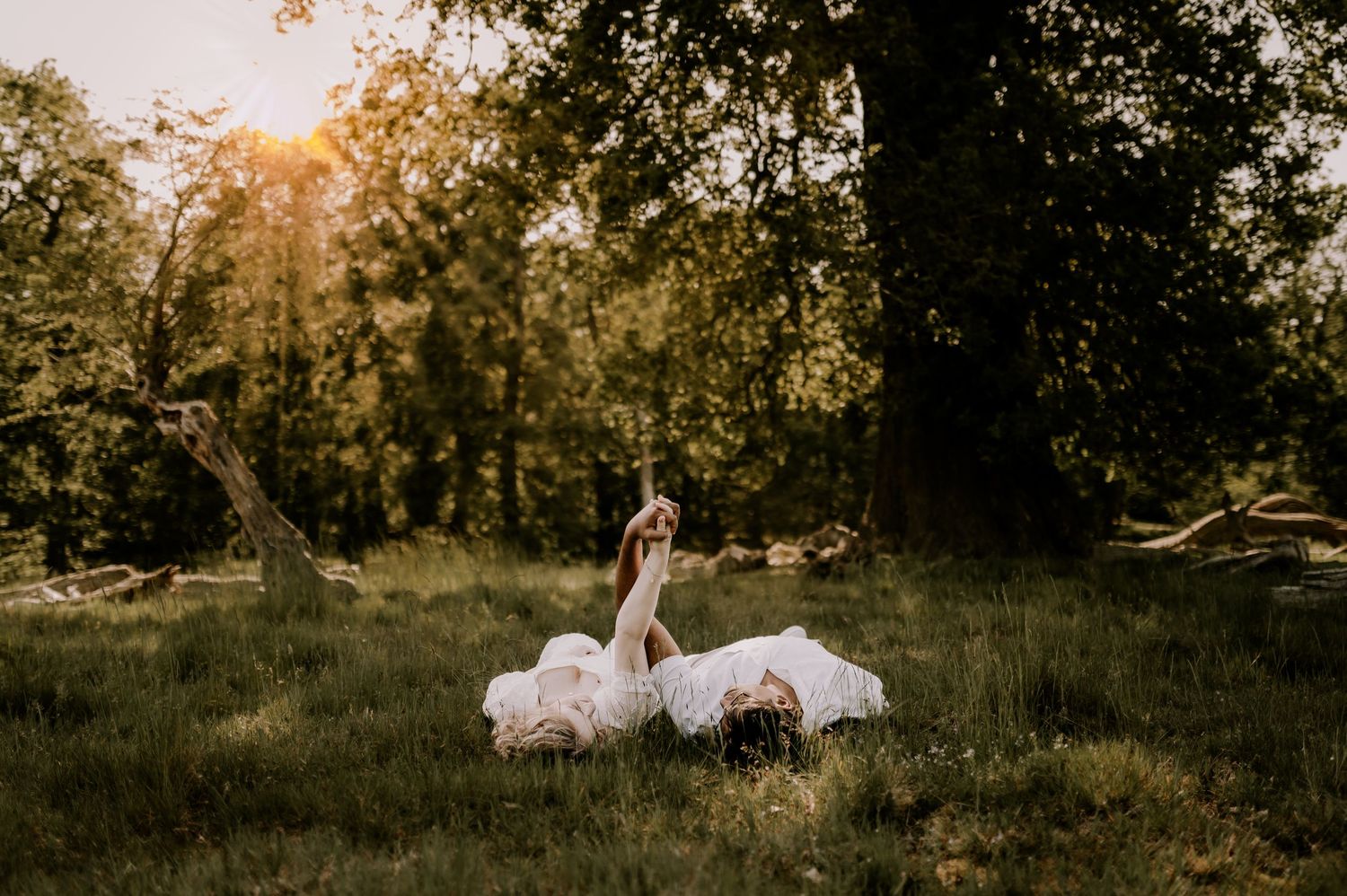 A bride in a white dress and groom lie on lush green grass in golden hour sunlight beneath tall trees.