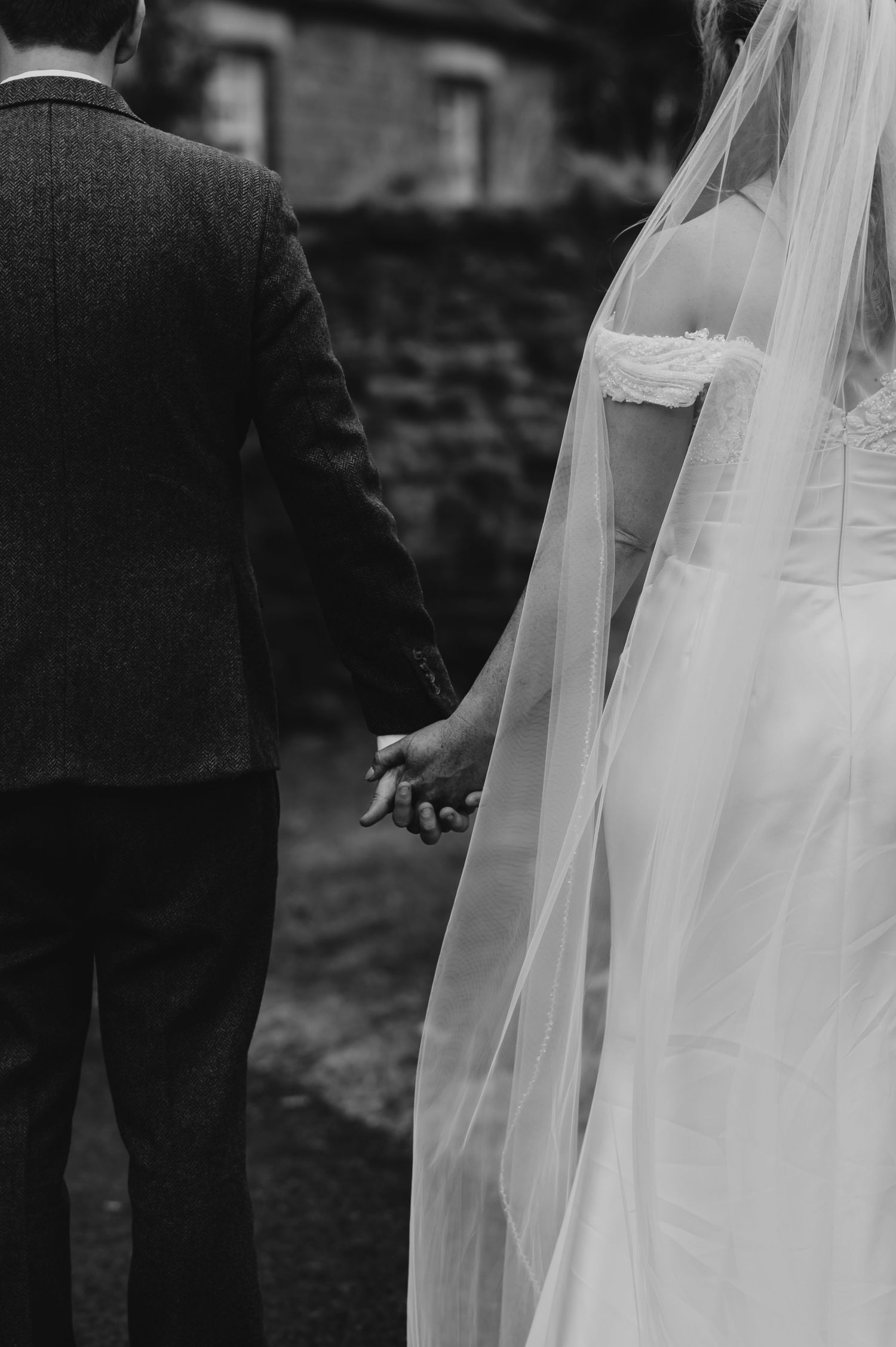 A black and white wedding photograph showing the flowing veil of a bride next to a suited figure outdoors.