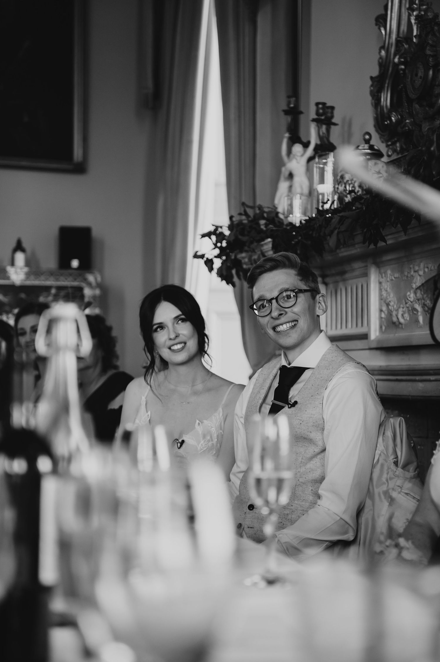 Black and white candid wedding reception photo showing guests seated at dinner table with candles in foreground.