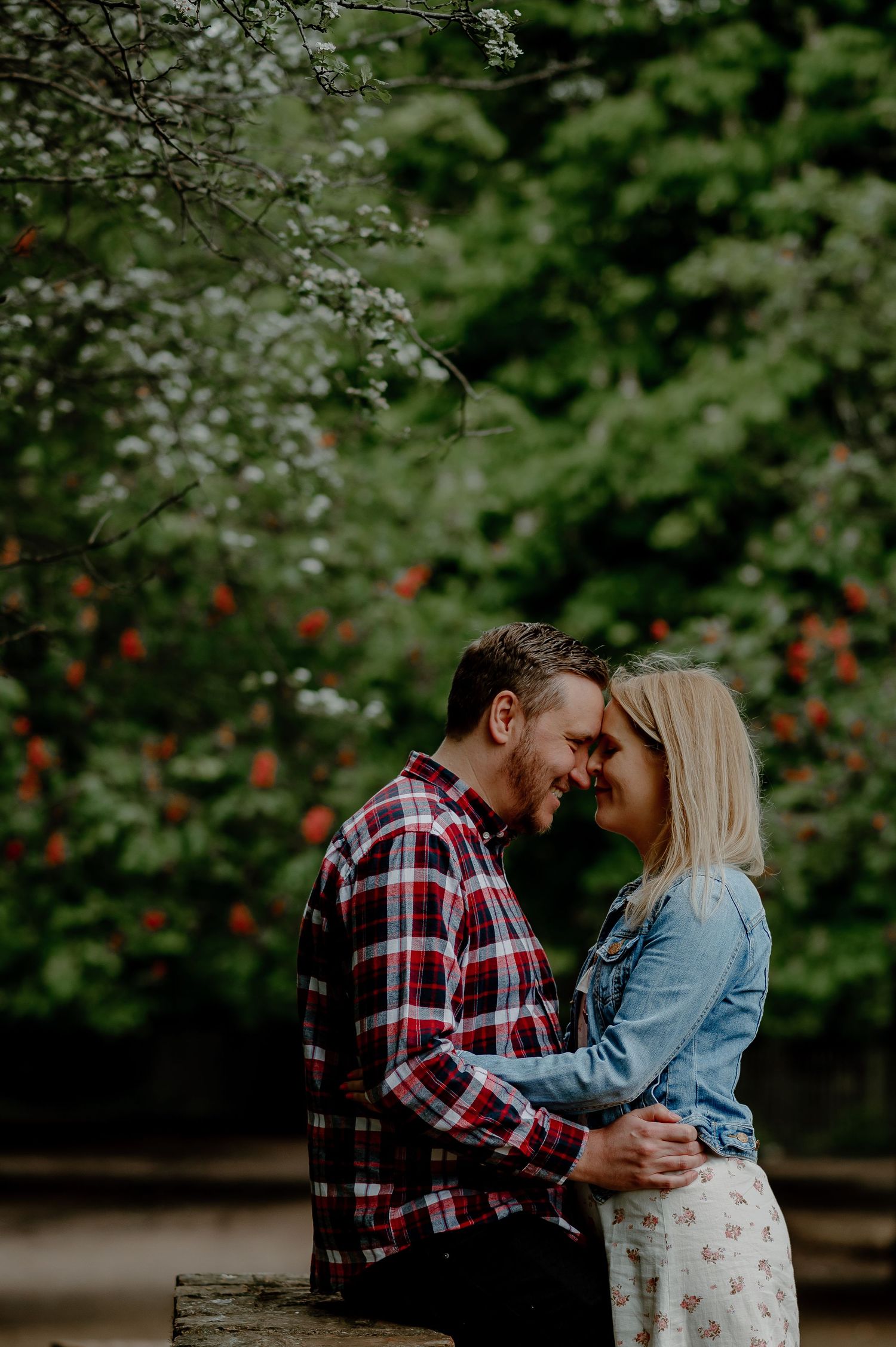 A couple shares an intimate moment in a garden setting surrounded by lush greenery and red flowers.
