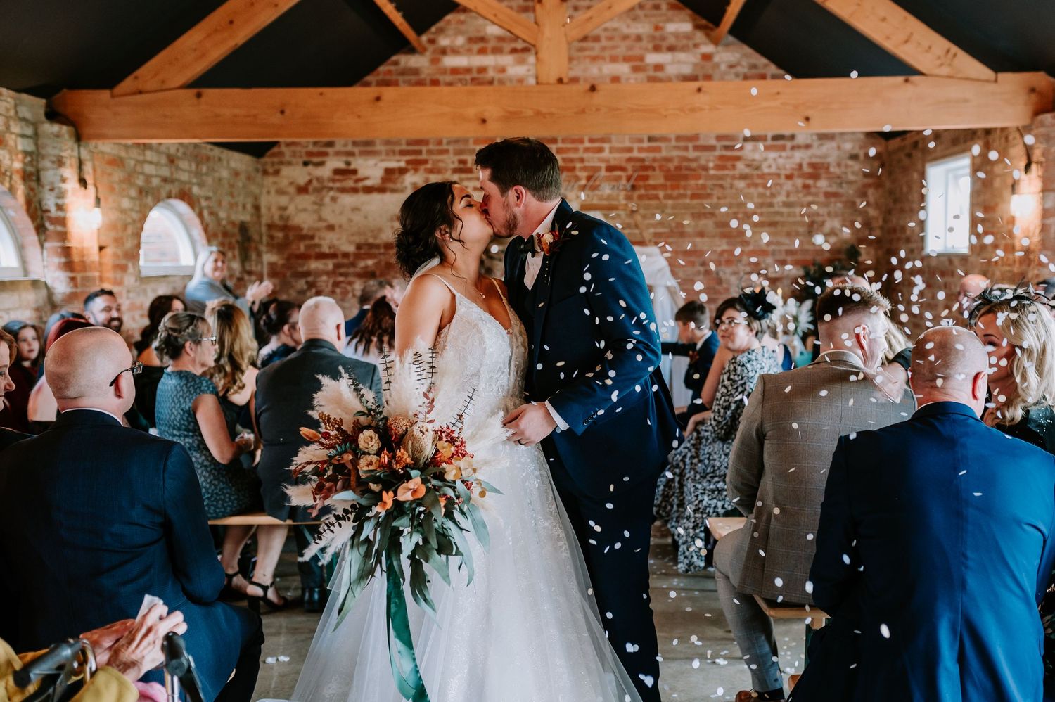 A romantic kiss at a rustic brick barn wedding venue as confetti falls around the celebrating couple.