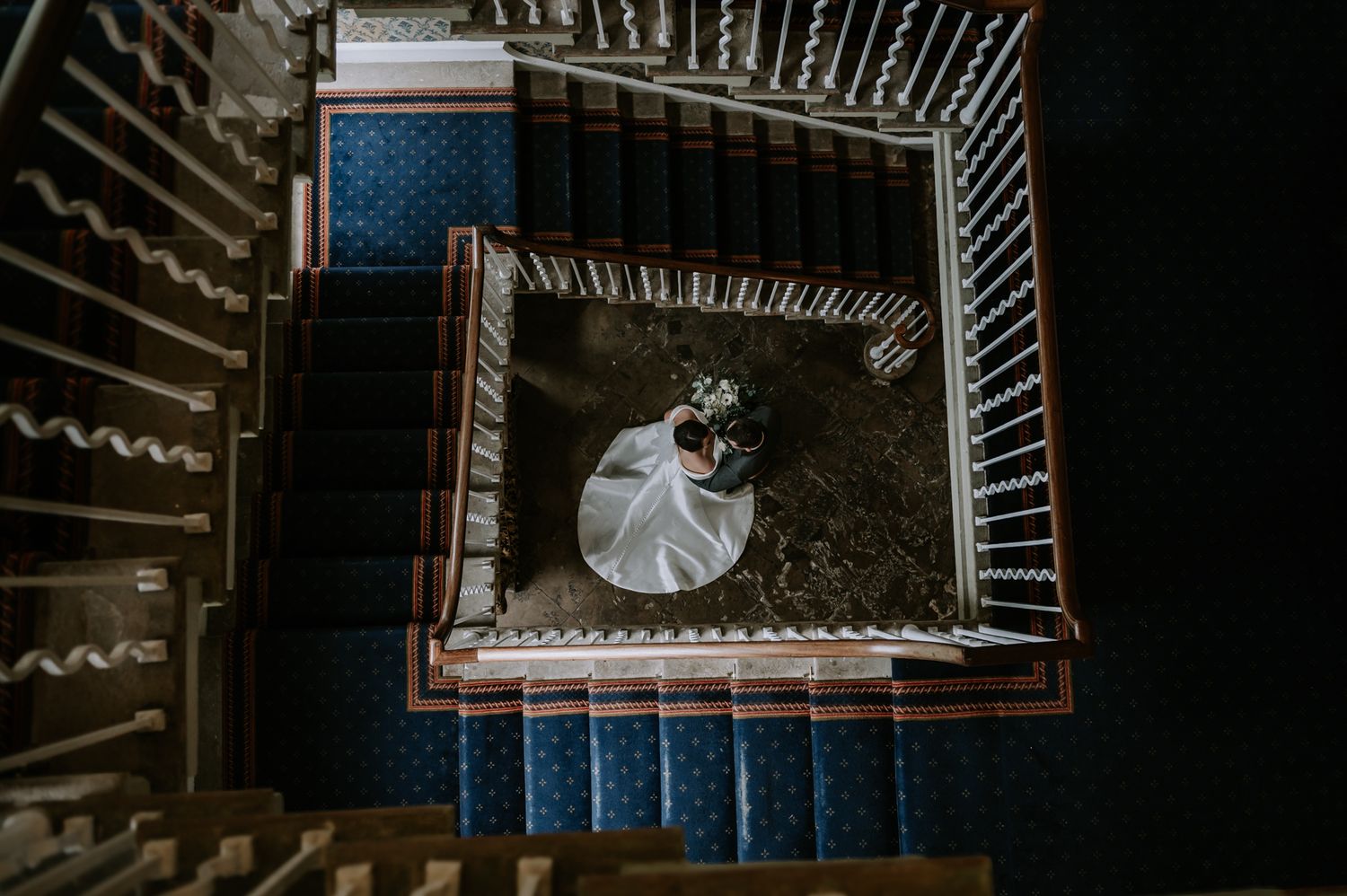 Overhead view of an elegant figure in a white dress descending a spiral staircase with ornate railings and blue carpet.