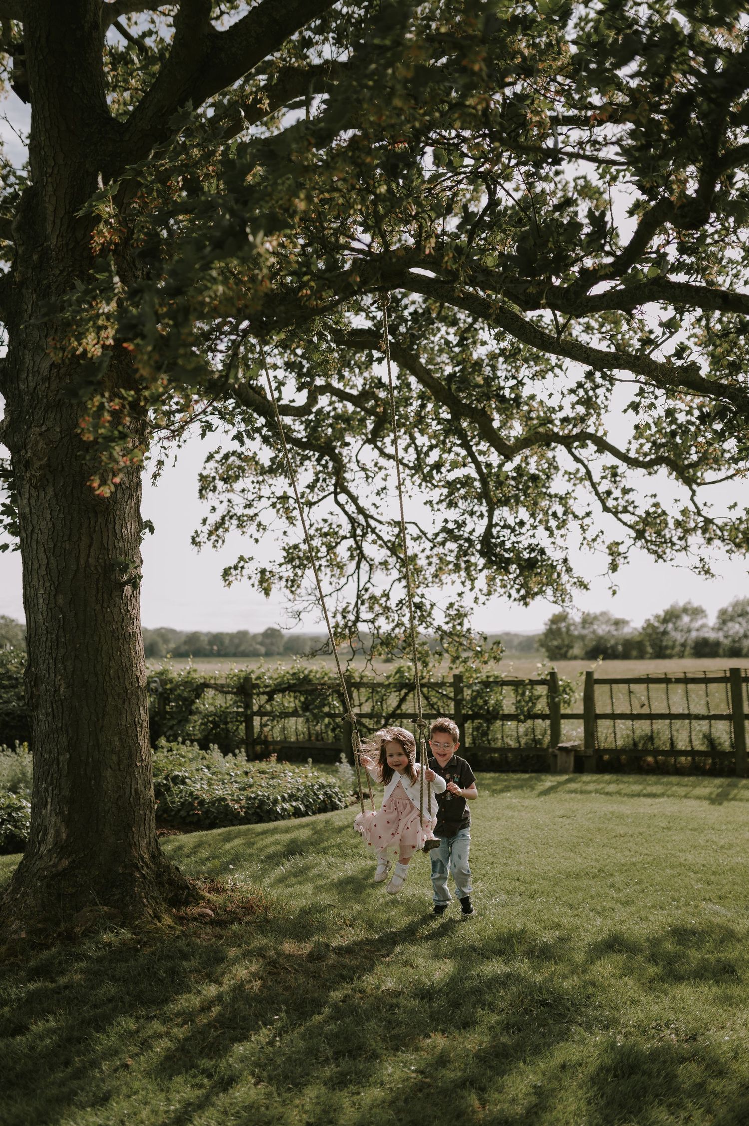 Two children playing on a rope swing hanging from a large oak tree in a lush green garden.