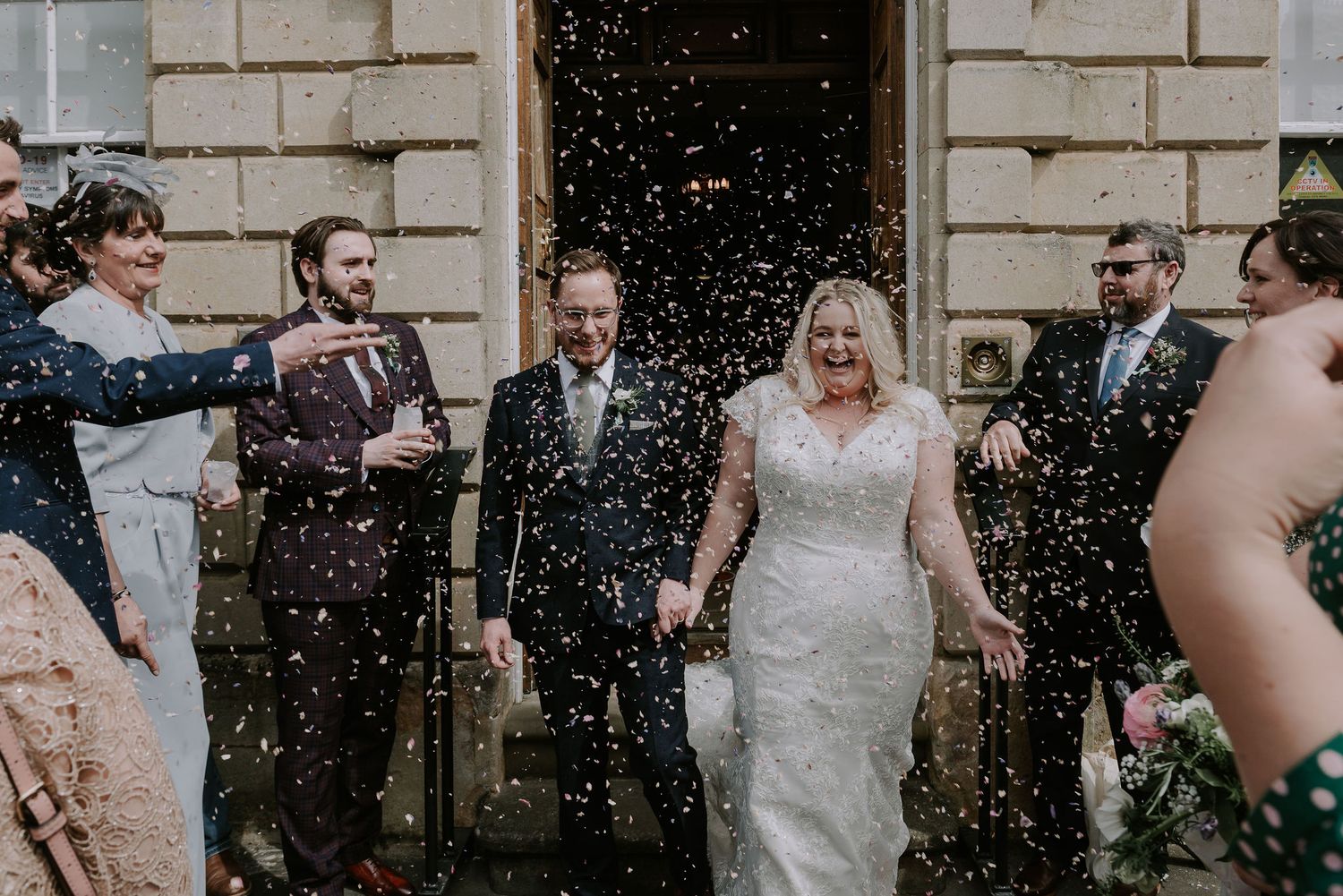 Wedding celebration outdoors with guests throwing confetti as newlyweds laugh in the middle
