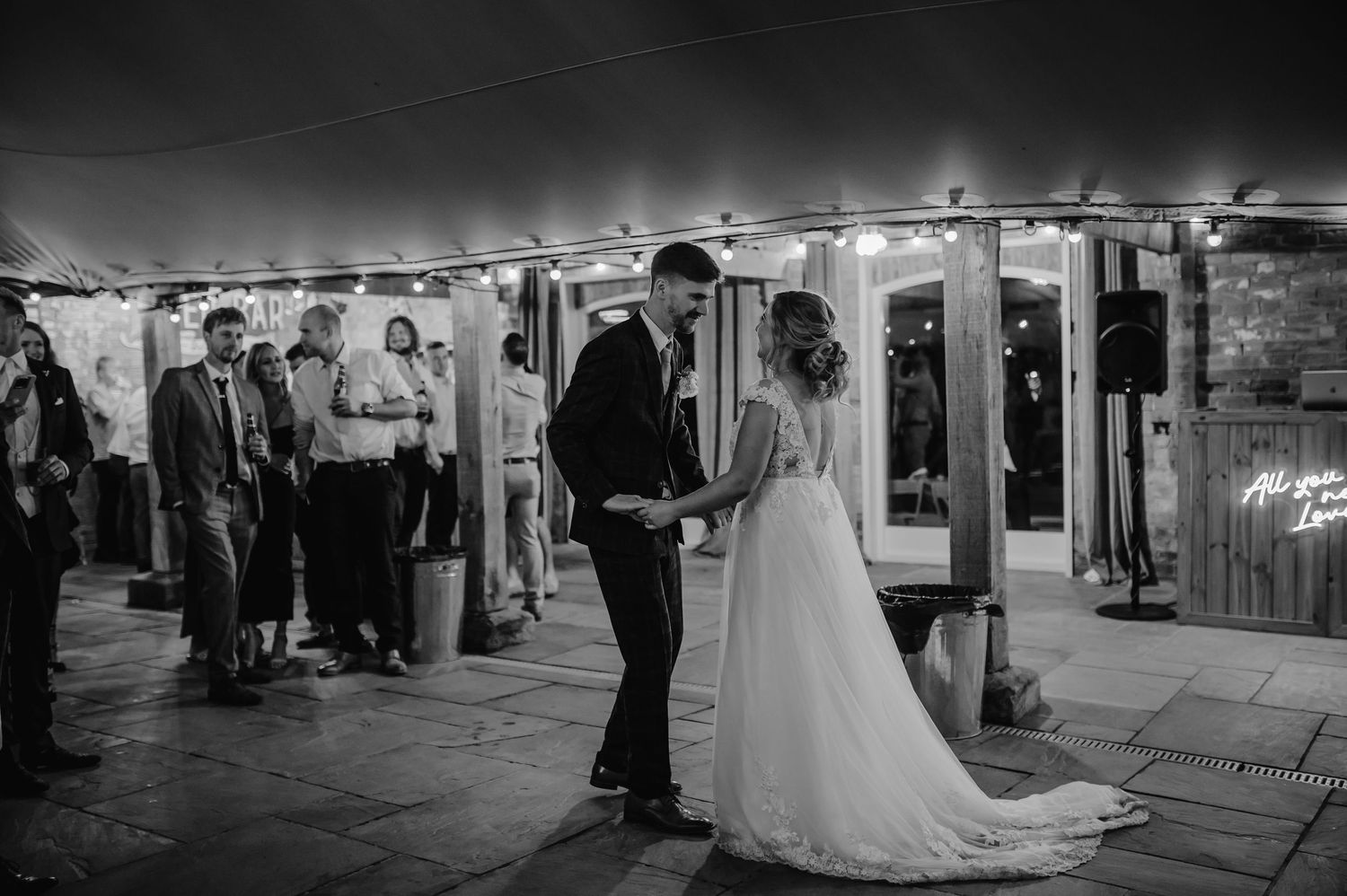 A couple stands together at their evening wedding reception under string lights in a stone courtyard.